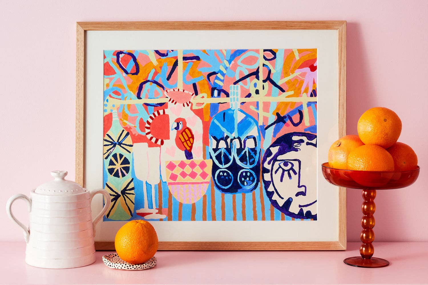 Colorful abstract art print in a wooden frame on a pink wall with oranges and a teapot.