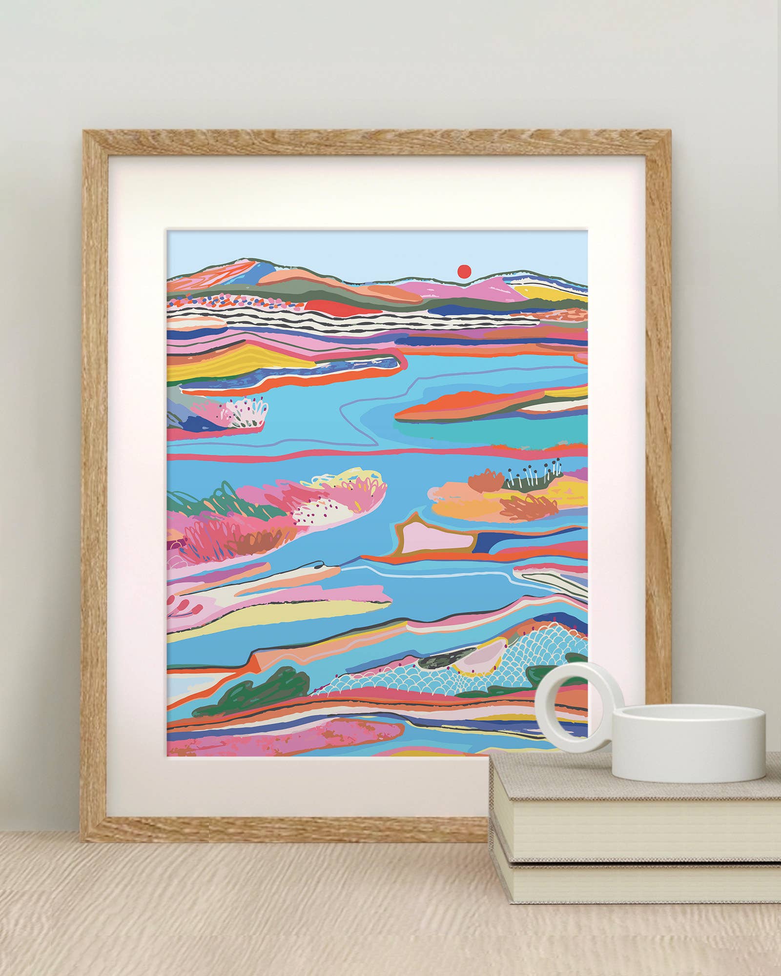 Framed colorful abstract landscape print on a wall with a cup and books below.