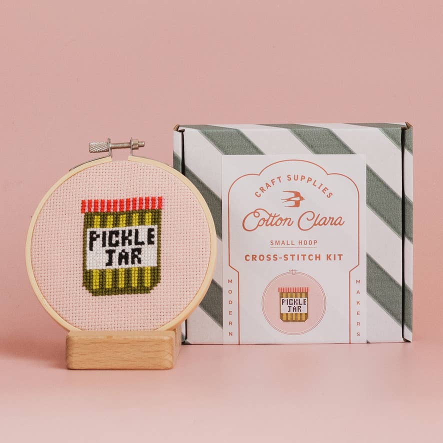 Cross-stitch hoop with 'Pickle Jar' design next to Cotton Clara cross-stitch kit box on a pink background