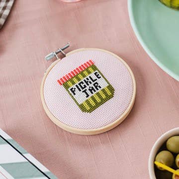 Cross-stitched hoop with 'Pickle Jar' design on a pink surface