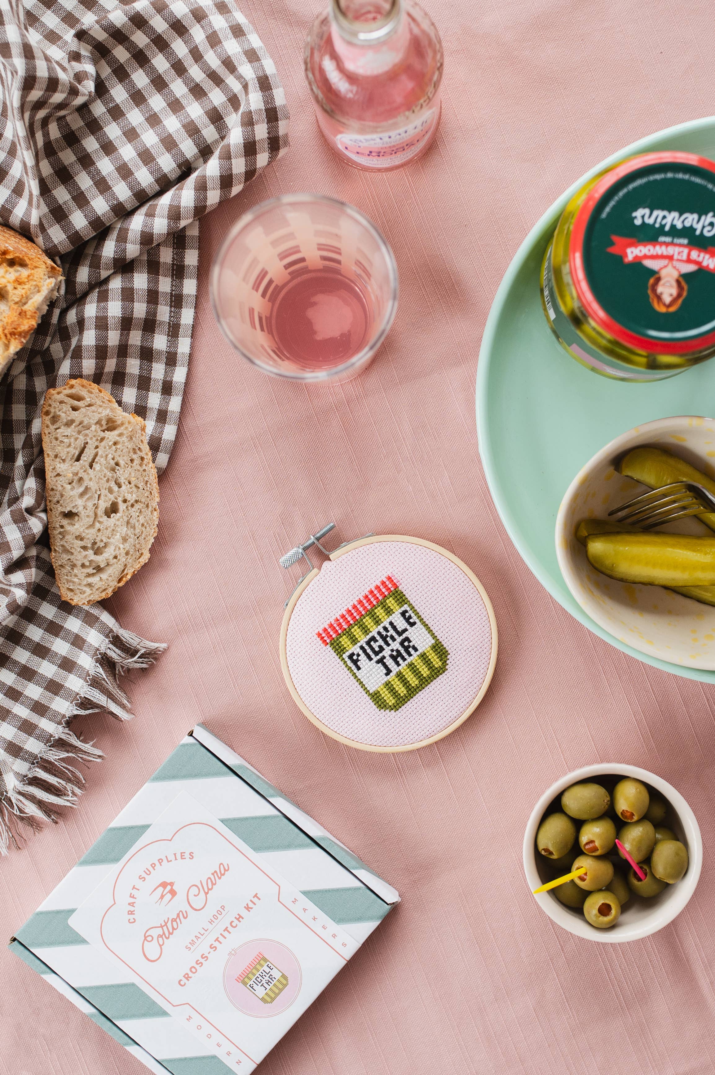 Table setting with bread, pickles, and a pickle-themed embroidery hoop on a pink surface.