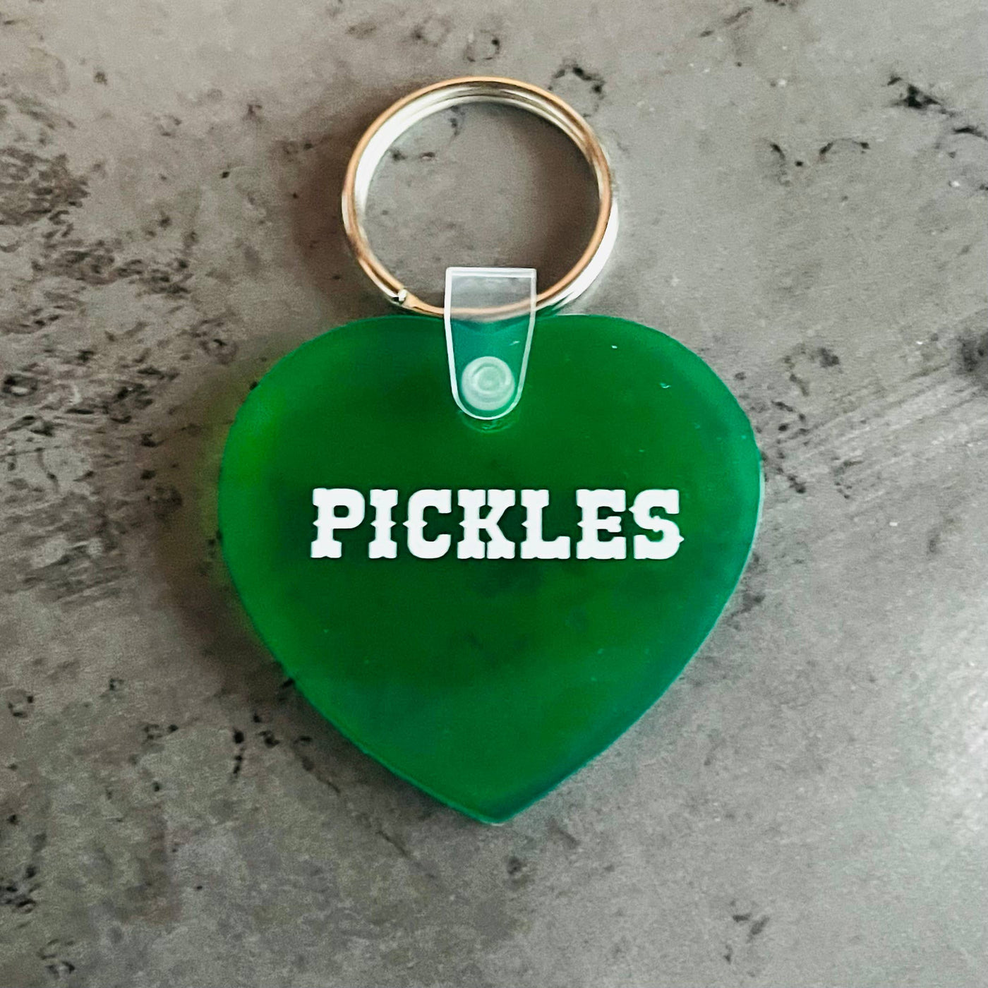 Green heart-shaped keychain with 'PICKLES' text on a concrete surface