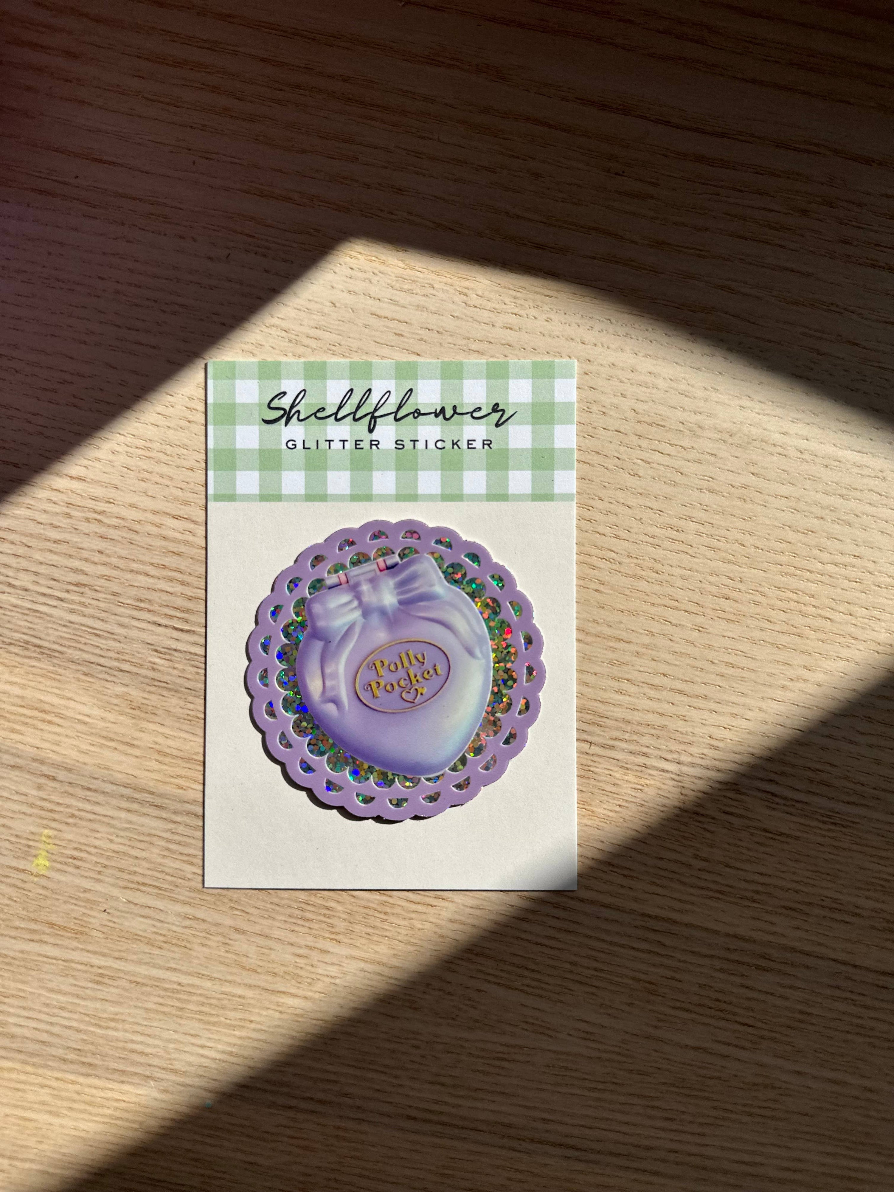 Purple glitter sticker on a card with 'Shellflower' branding on a wooden surface