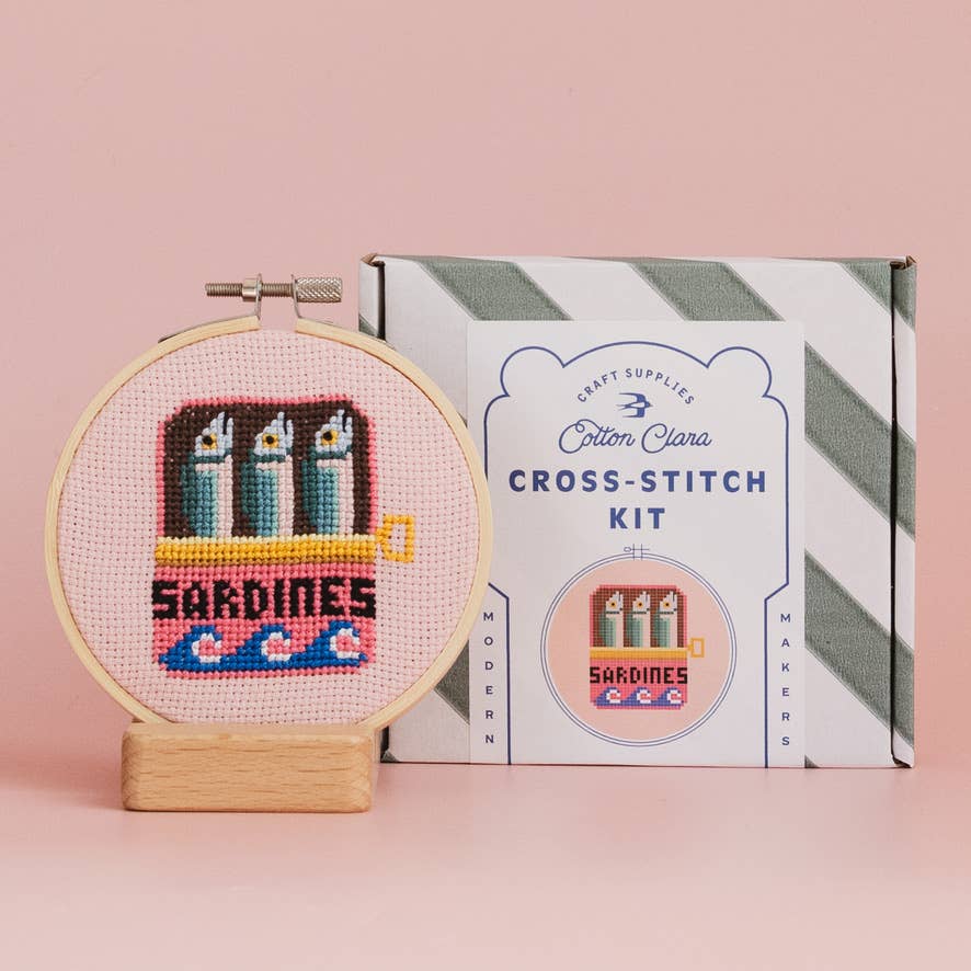 Cross-stitch kit with completed design of sardines in a can on a pink background