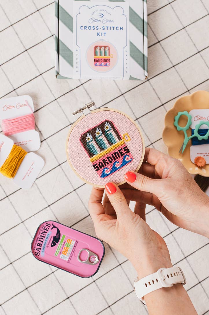 Person holding a cross-stitch kit with 'Sardines' design on a tiled surface.