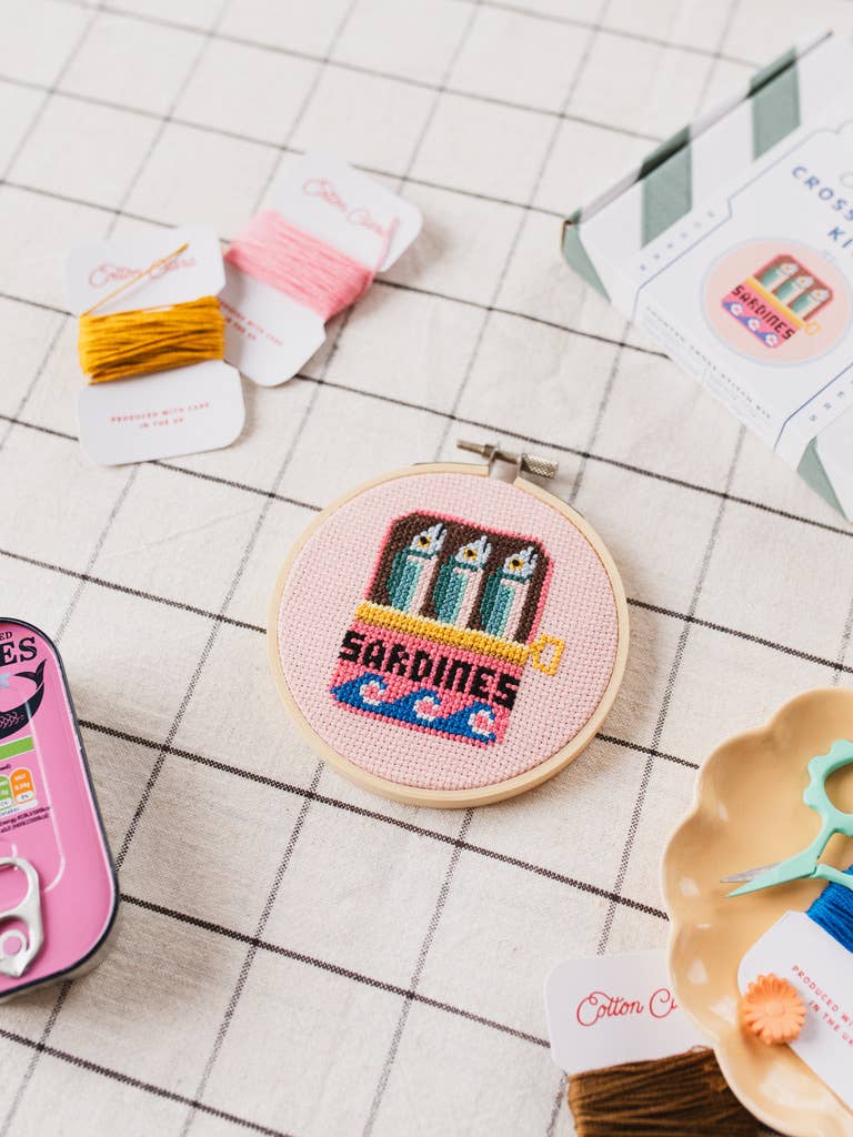 Embroidery hoop with 'Sardines' design on a grid background with embroidery supplies.