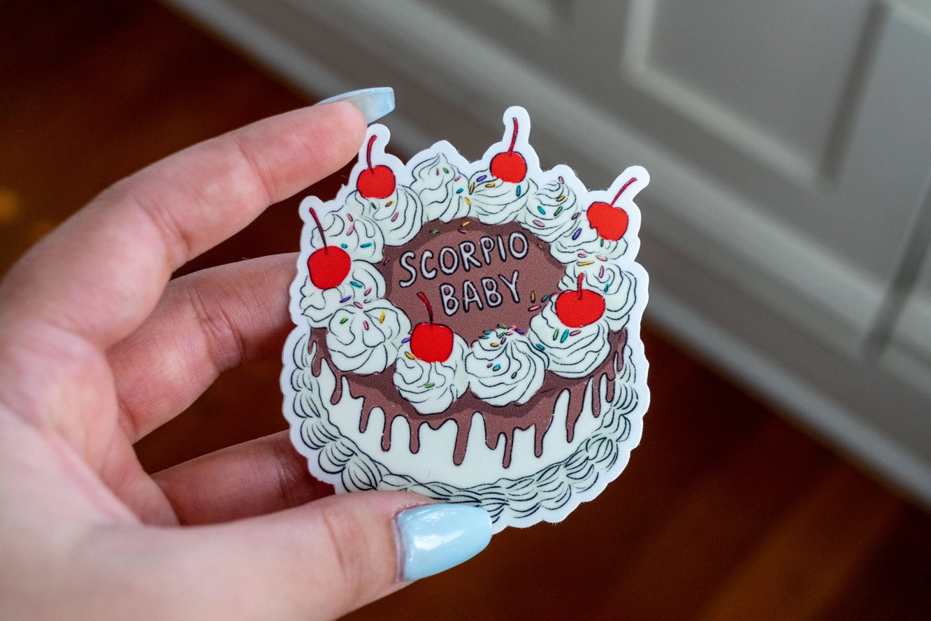 Hand holding a sticker shaped like a cake with 'Scorpio Baby' text.