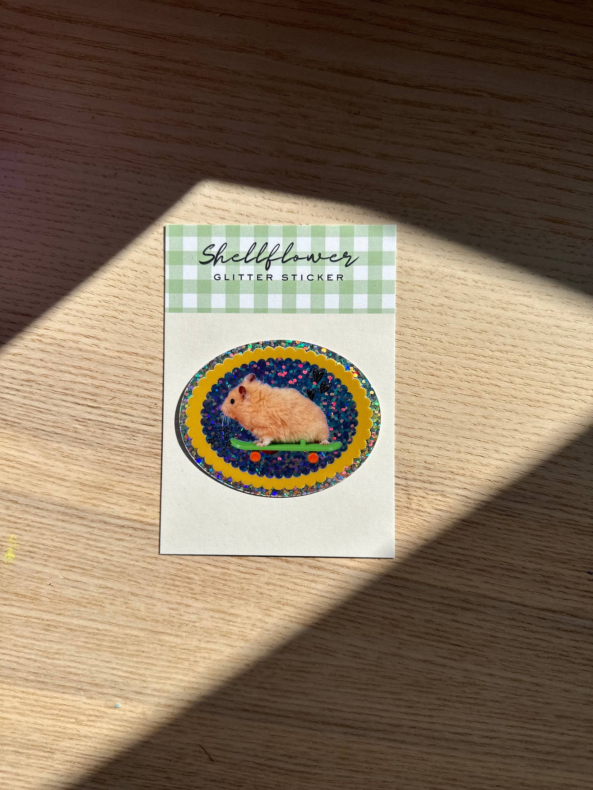 Hamster sticker on a Shellflower card with a checkered border, placed on a wooden surface.