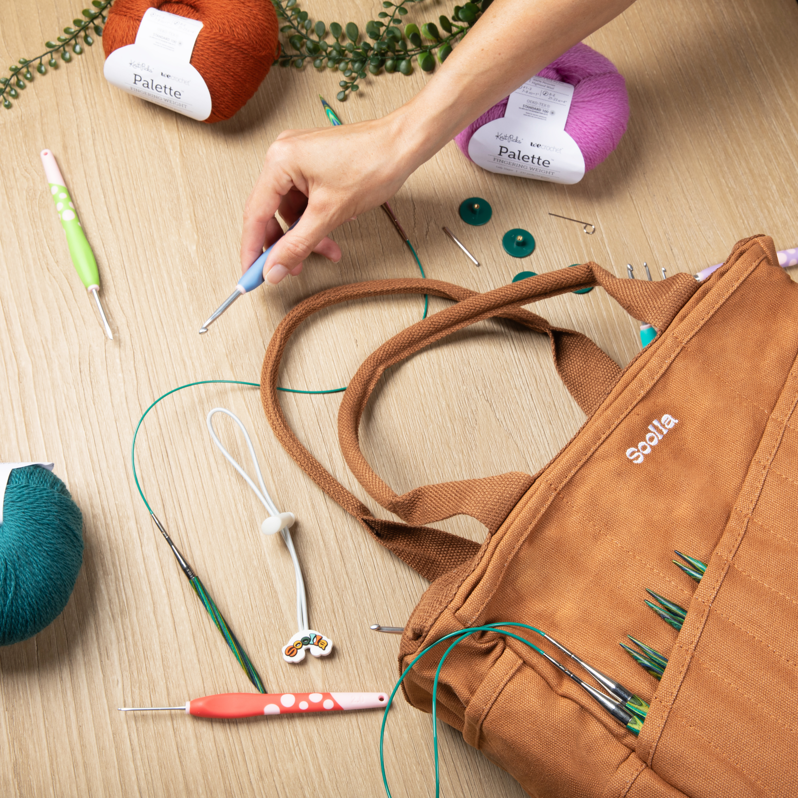 Person working with knitting needles and yarn on a wooden surface, with a brown bag containing needles.