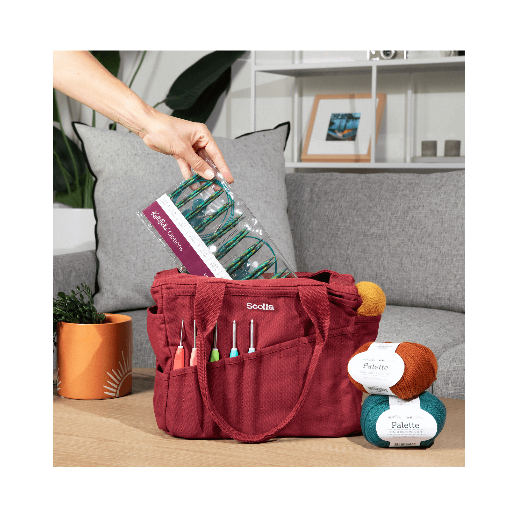 Red craft bag with knitting supplies on a wooden table, with a gray couch and plant in the background.