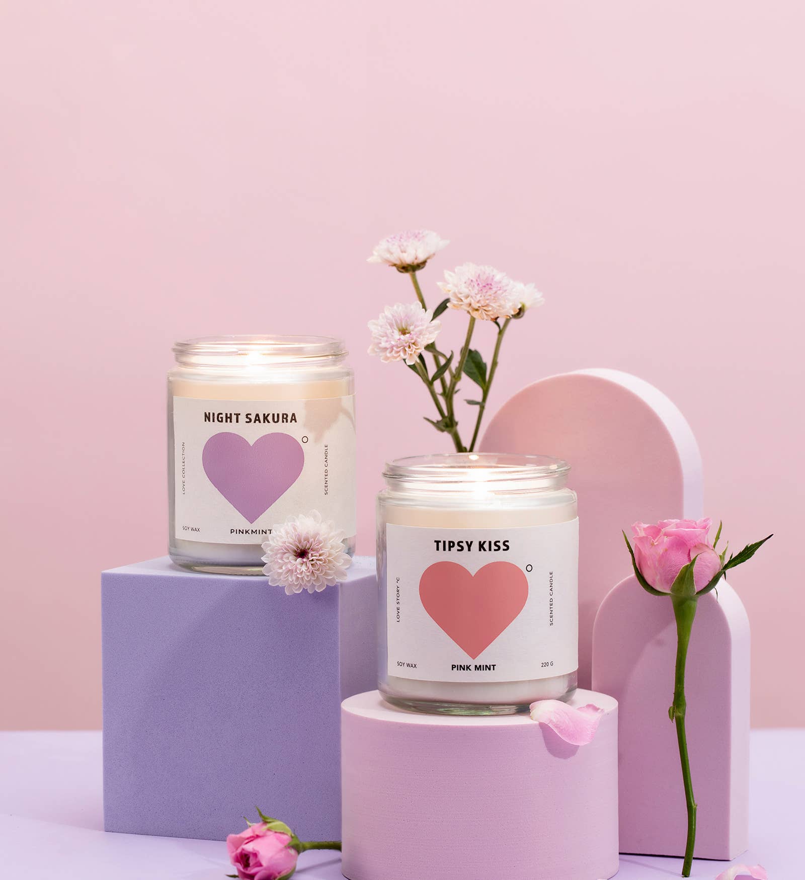 Two candles with heart designs on labels, surrounded by flowers against a pink background