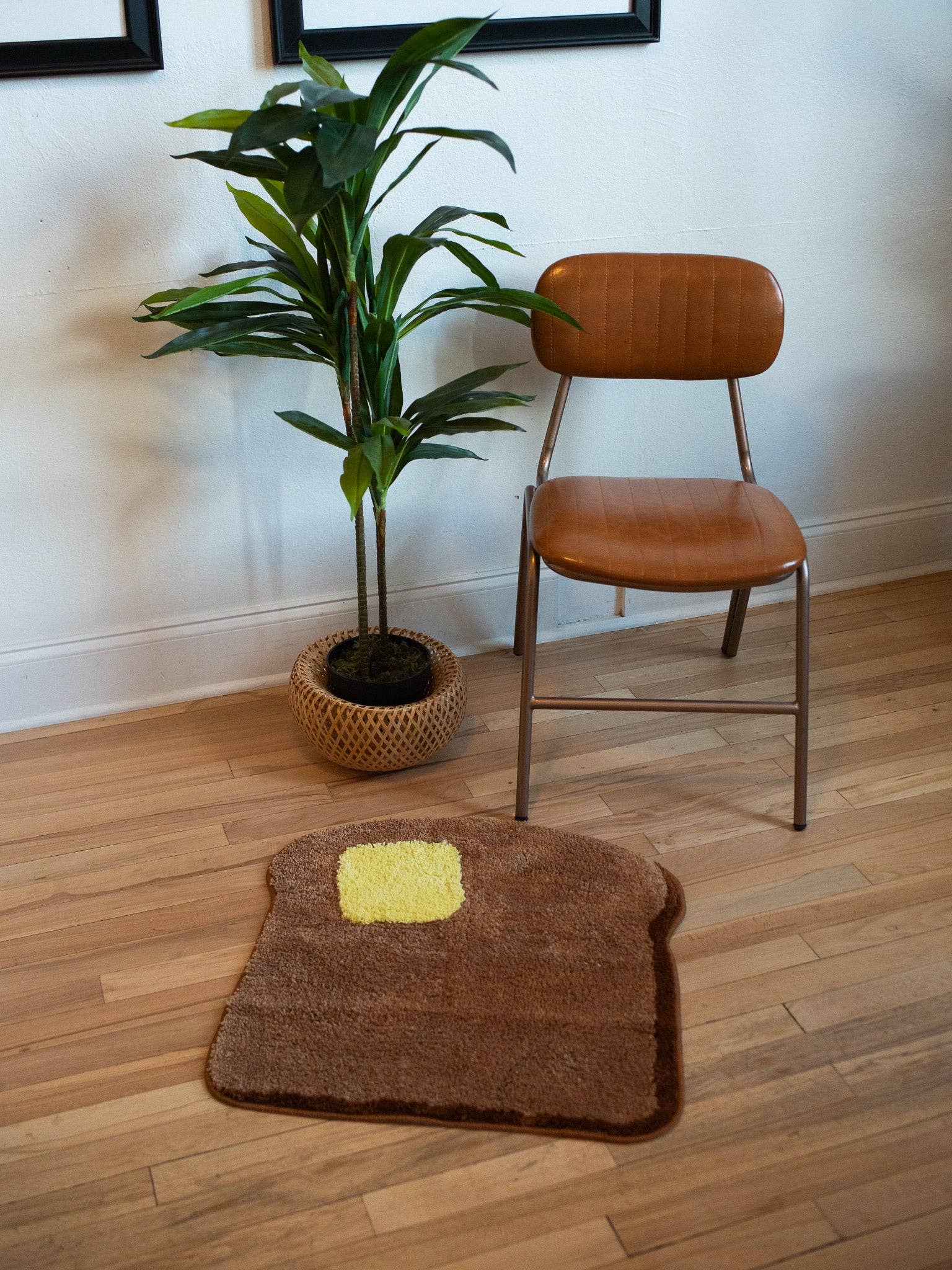 Brown rug shaped like a piece of toast with a yellow square on a wooden floor, next to a plant and a brown chair.