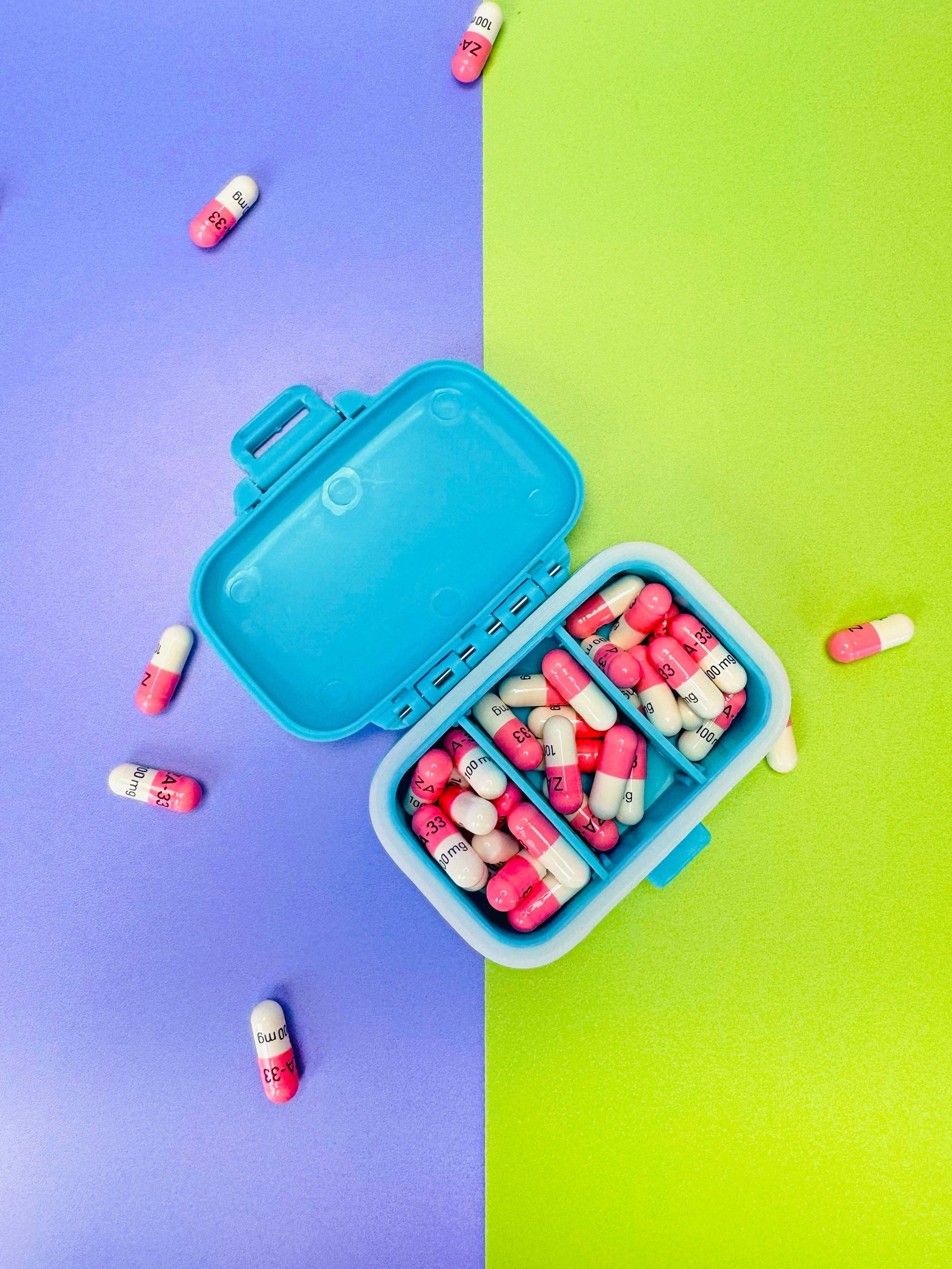 Blue pill organizer with pink and white capsules on a two-tone purple and green background