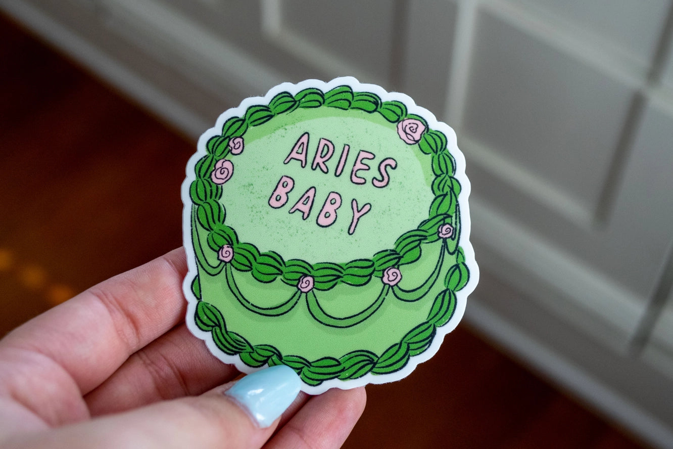 Hand holding a green sticker with 'ARIES BABY' text against a neutral background