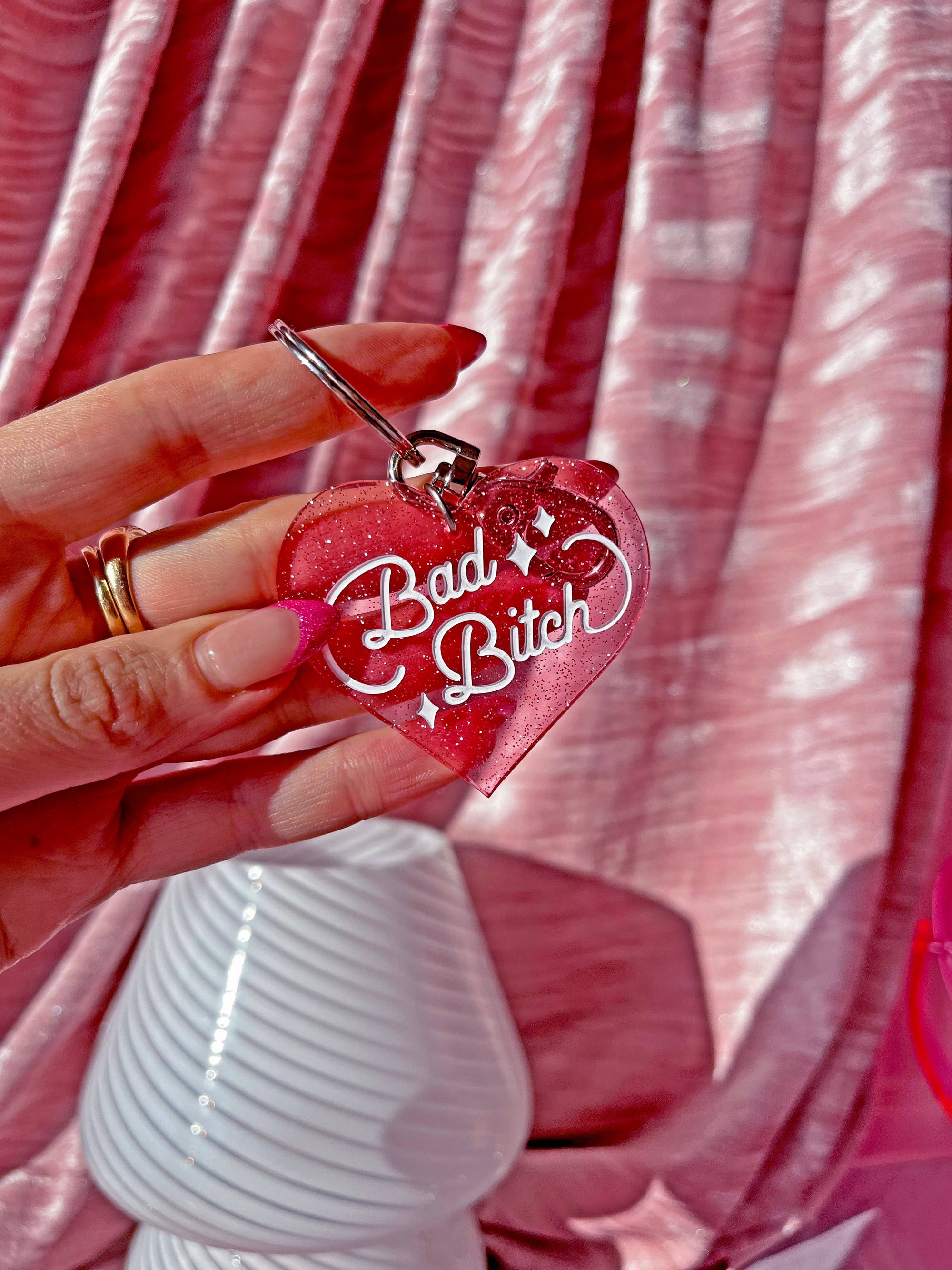 Heart-shaped keychain with 'Bad Bitch' text held by a hand against a pink background