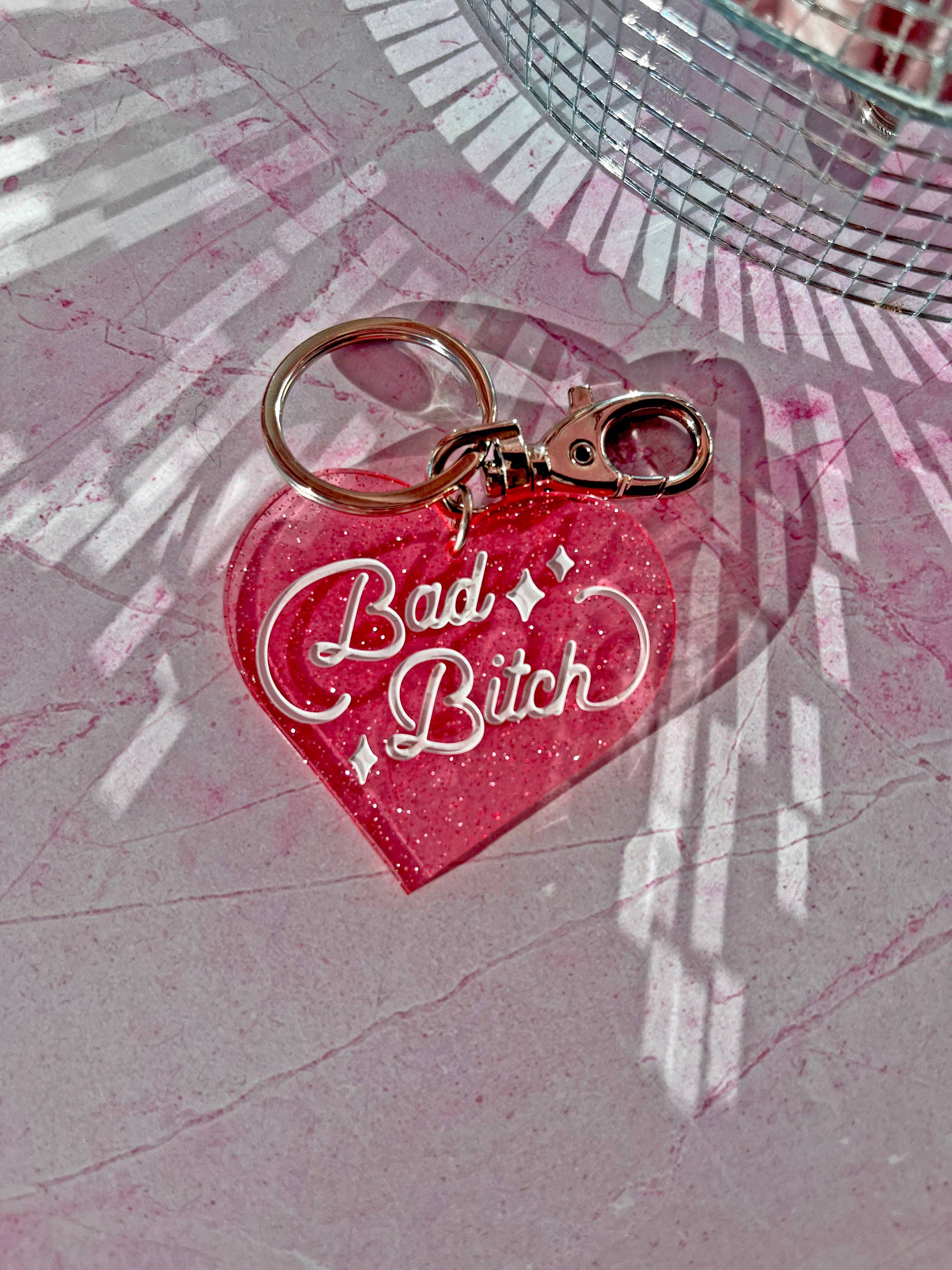 Heart-shaped keychain with 'Bad Bitch' text on a pink textured surface