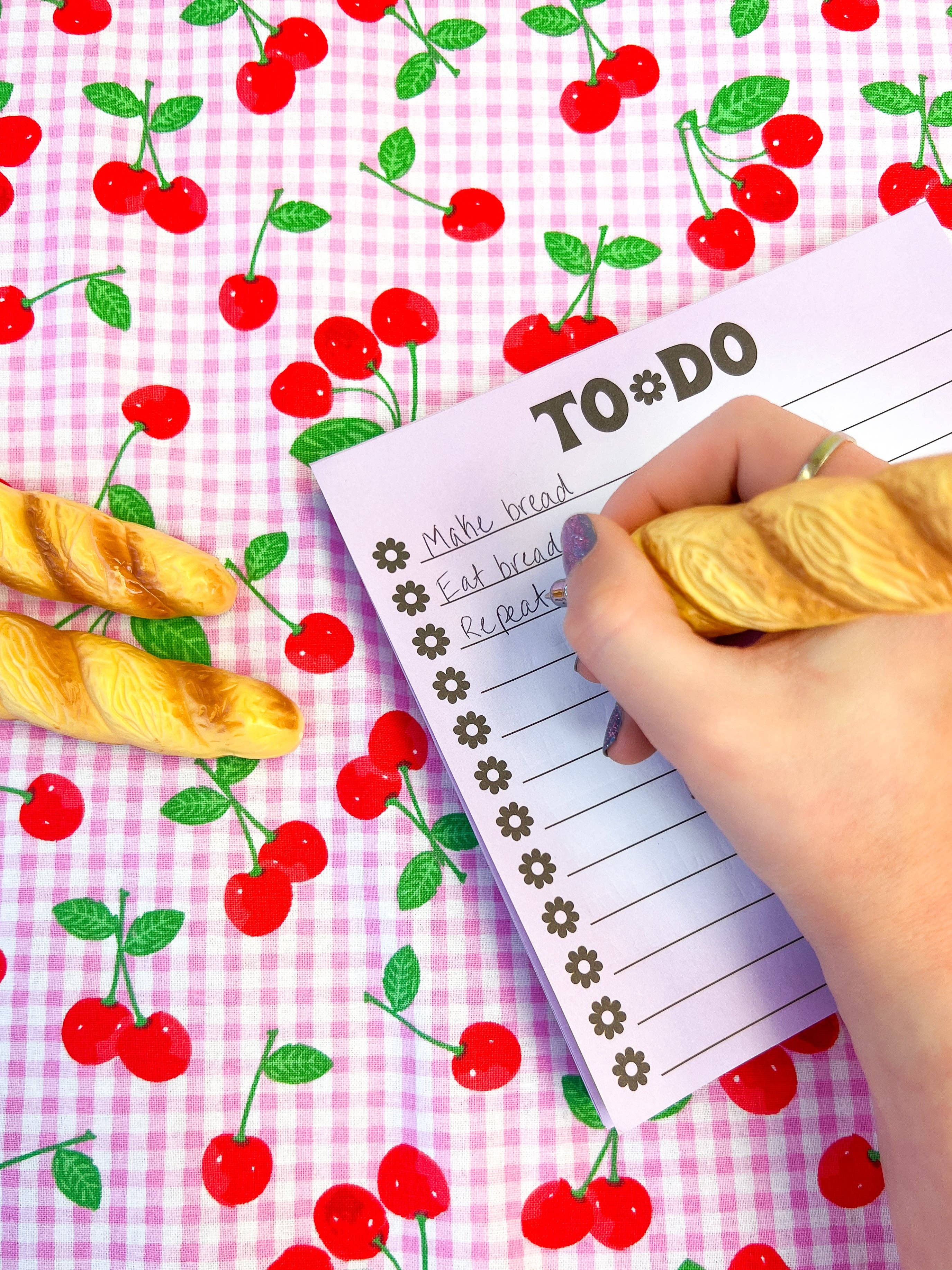 Hand writing on a to-do list with cherries and bread on a pink checkered background