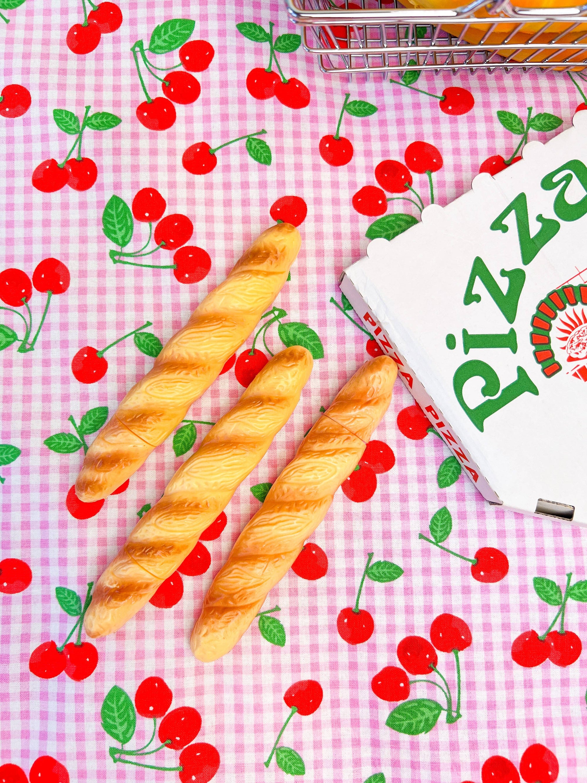 Three bread sticks on a pink checkered tablecloth with red cherries and a pizza box.