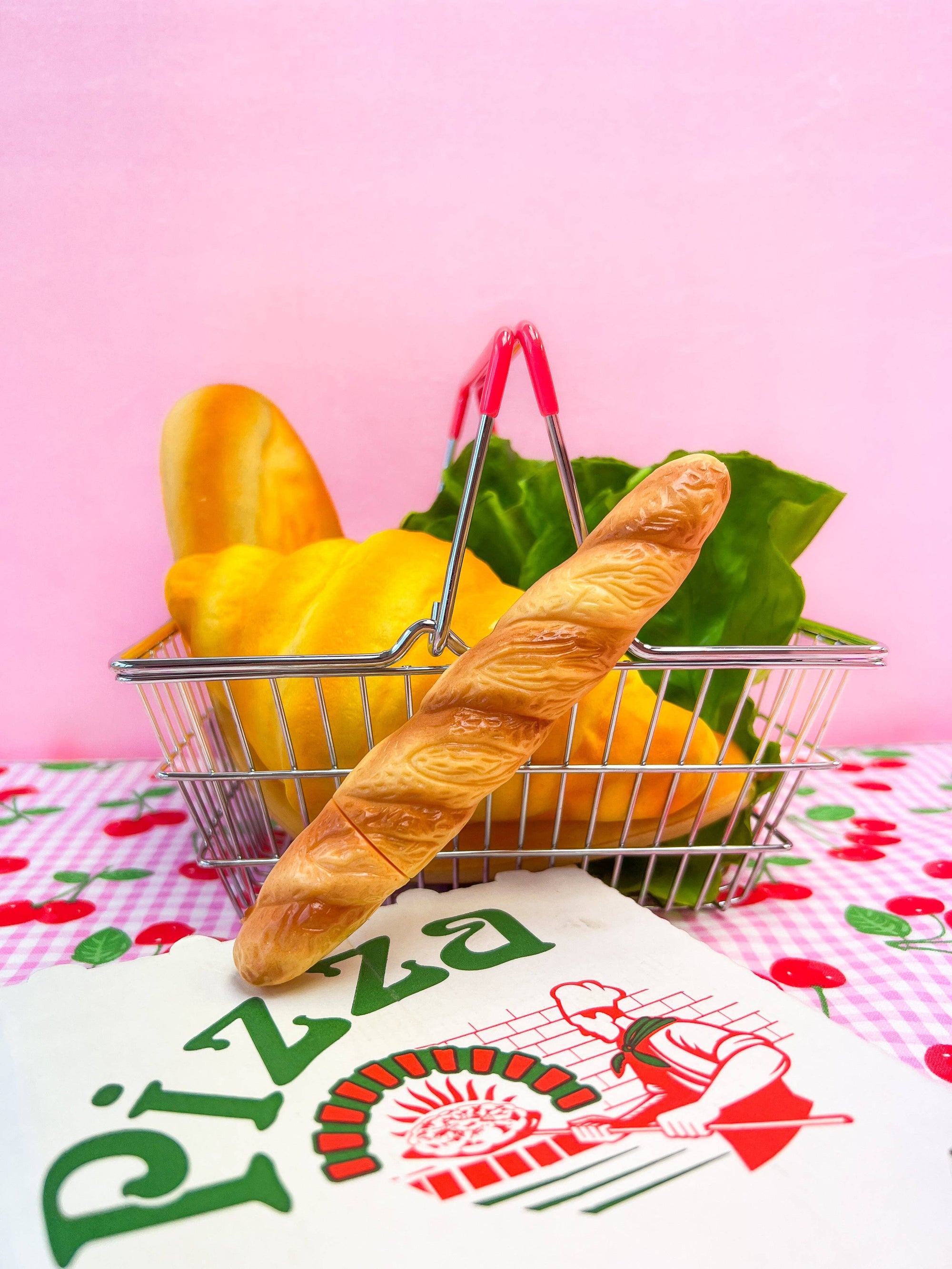 Metal shopping basket with bread, bananas, and lettuce on a pink background with a pizza-themed towel.