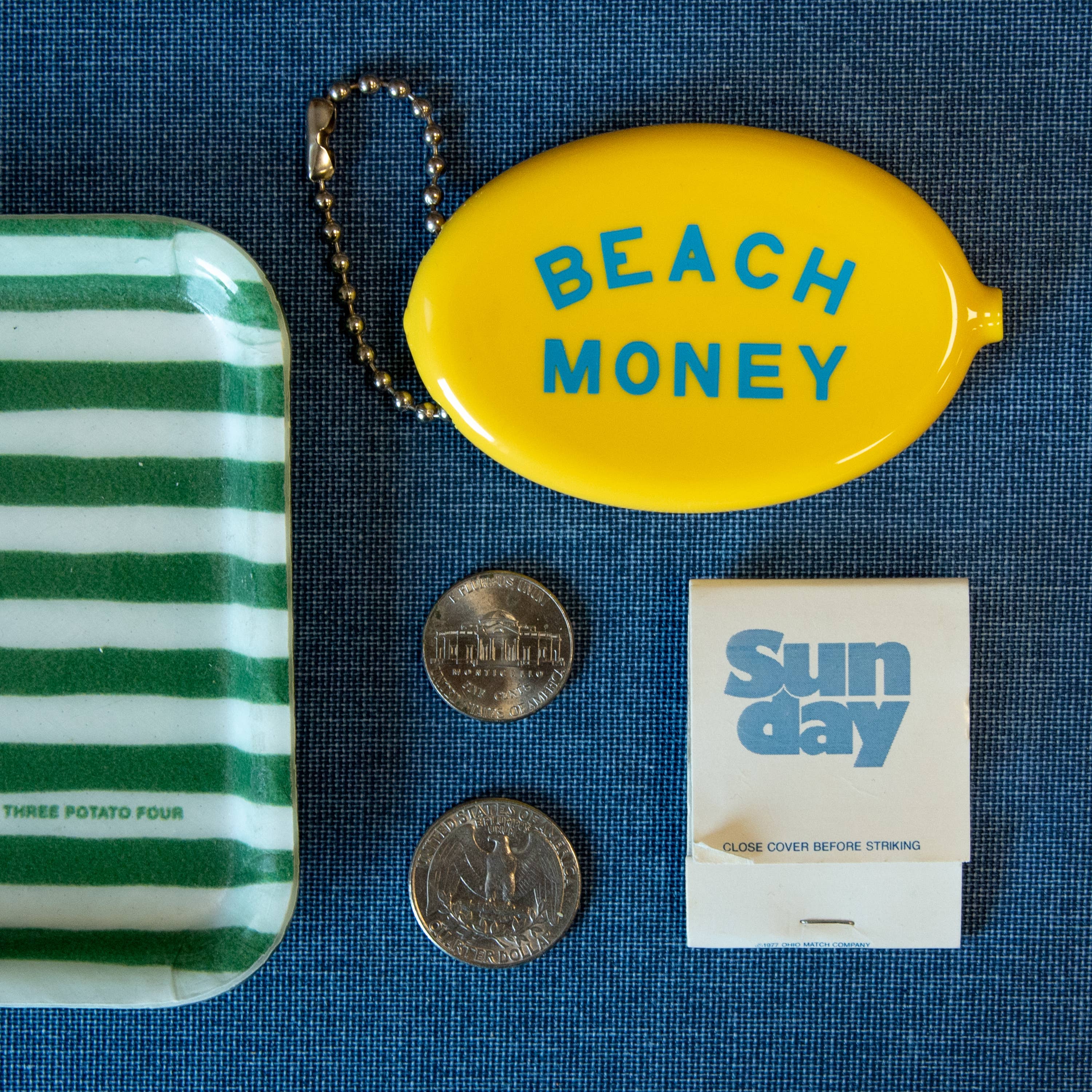 Collection of items including a green and white striped container, yellow 'Beach Money' pouch, coins, and a matchbook on a blue textured background.