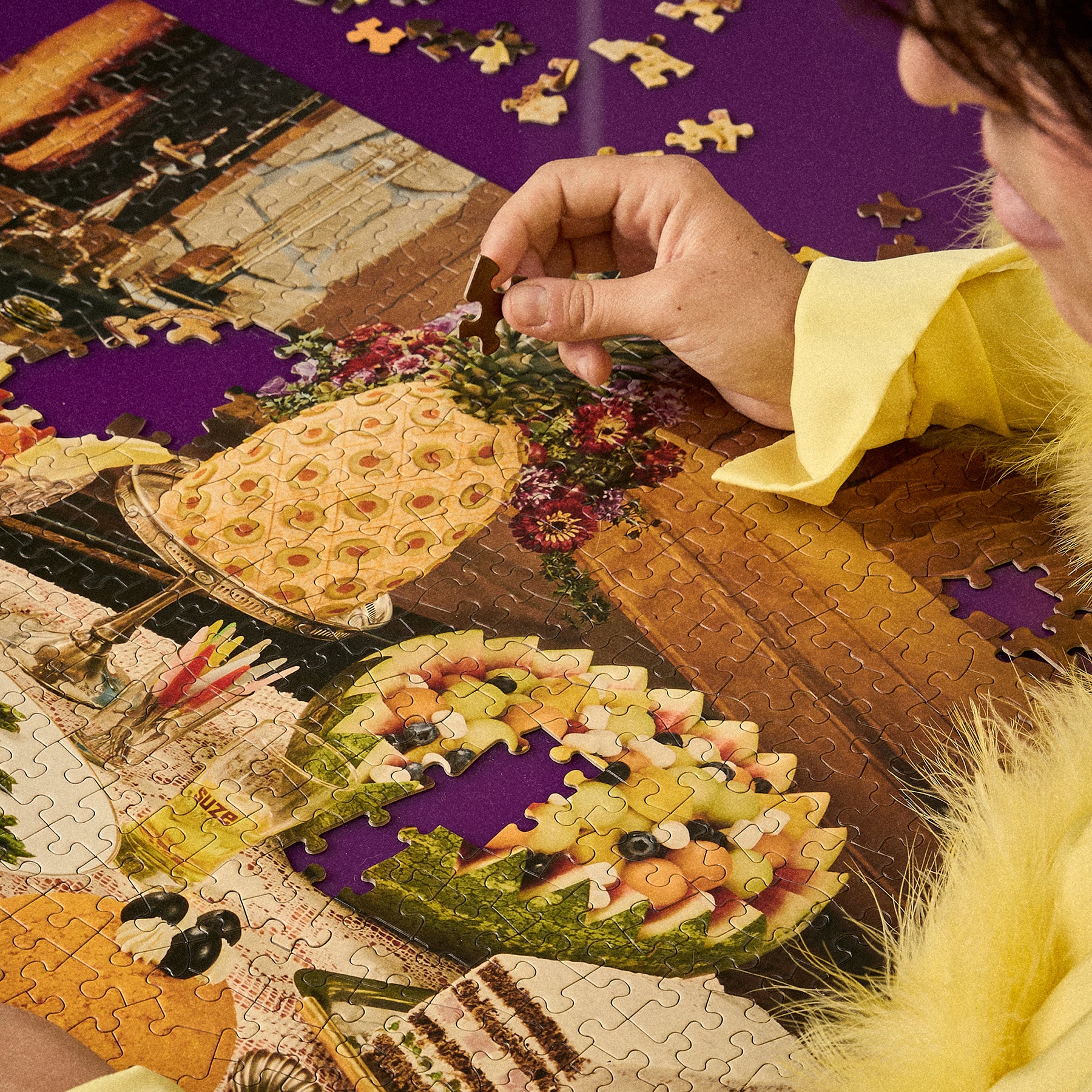 A close-up of a person assembling a colorful jigsaw puzzle featuring a dessert scene, with a hand placing a piece on a purple background and loose puzzle pieces scattered around.