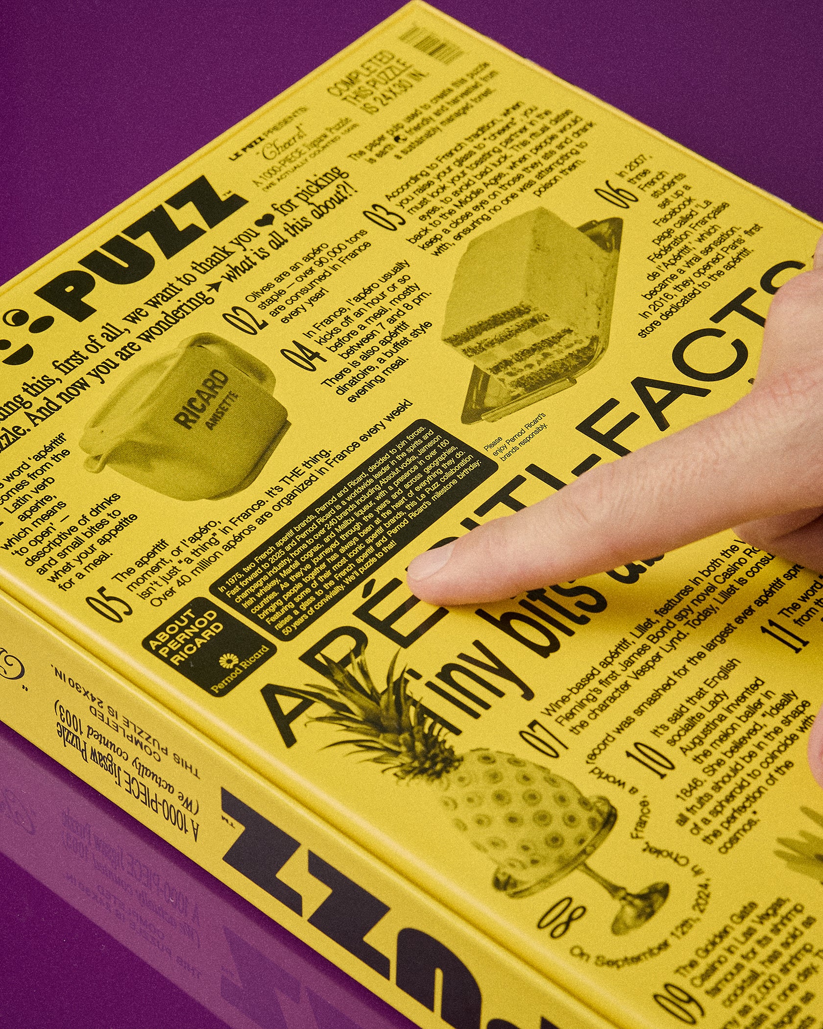 A close-up of a yellow 'Le Puzz' mini puzzle book being held by a person's hand, with the finger pointing at text on the cover against a purple background.