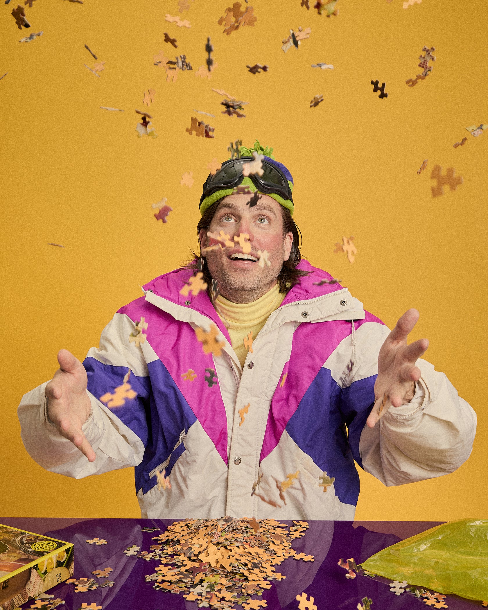 A man wearing a colorful vintage-style jacket, yellow turtleneck, and winter hat with goggles stands with his hands outstretched as autumn leaves and jigsaw puzzle pieces fall around him against a solid yellow background.