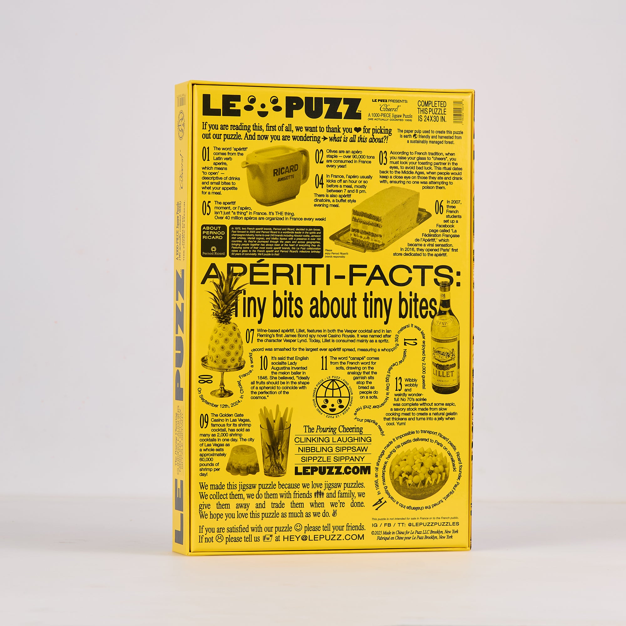 A yellow rectangular printed poster or booklet titled 'LE PUZZ Aperiti-Facts: Tiny bits about tiny bites' stands upright against a light gray studio background. The design is mostly yellow with black text and small black illustrations, including a bottle, pineapple, bread, and a globe.