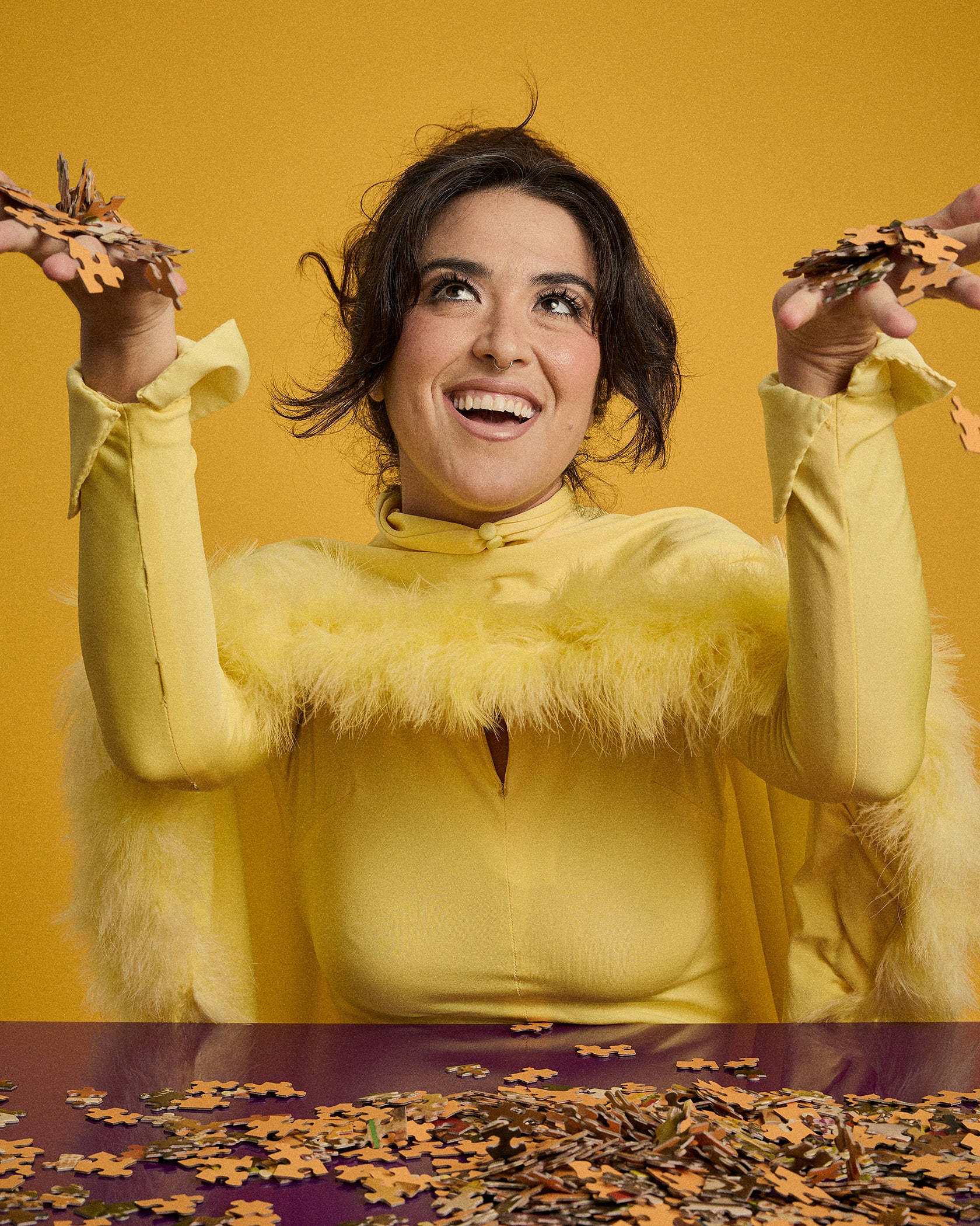 A woman with dark hair smiles while looking upward, holding scattered autumn leaves in both hands against a solid mustard-yellow background. She is wearing a bright yellow long-sleeve top with a fluffy faux-fur collar.
