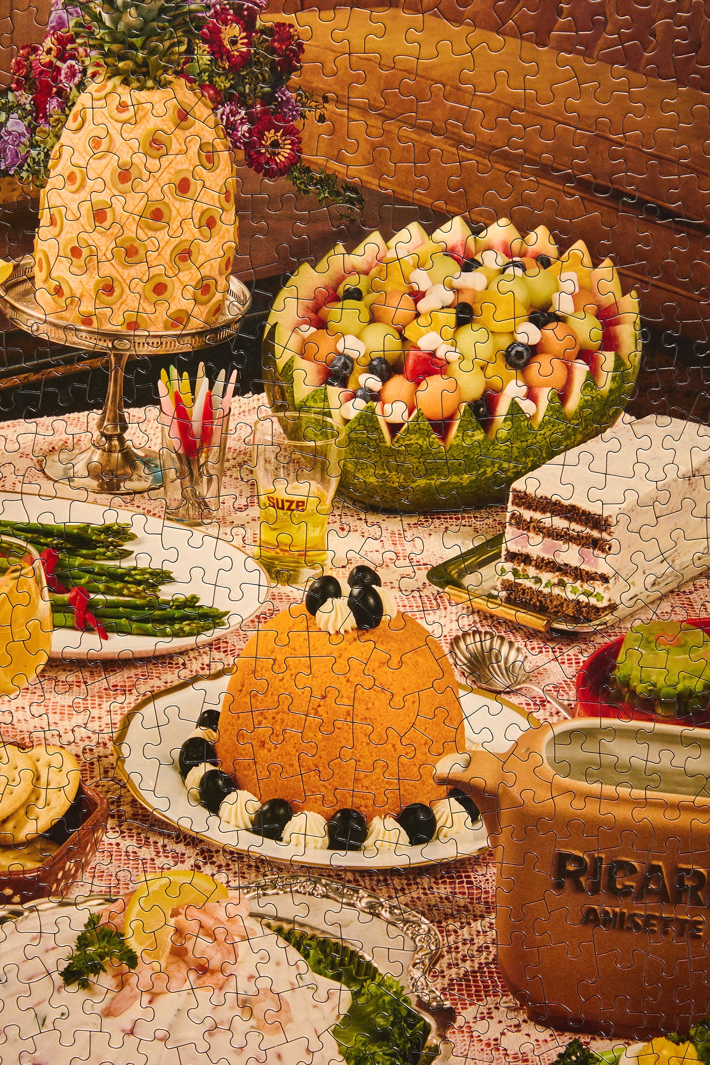 A completed jigsaw puzzle showing a festive table spread with a pineapple cake, fruit salad in a bowl, an orange mound dessert, asparagus, a slice of layered cake, and a brown ceramic mug with the text 'RICAR ANIKETTE'.