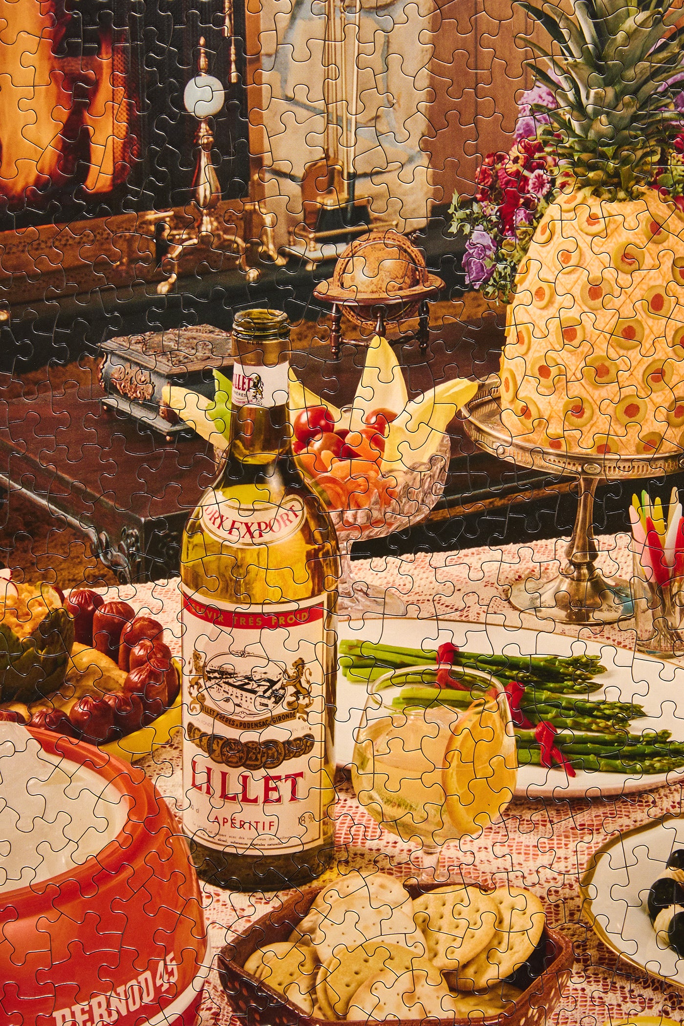 A completed jigsaw puzzle showing a festive table scene with a bottle of Lillet cognac, sliced lemons, crackers, asparagus, grapes, and a pineapple on a pedestal in a warmly lit interior.