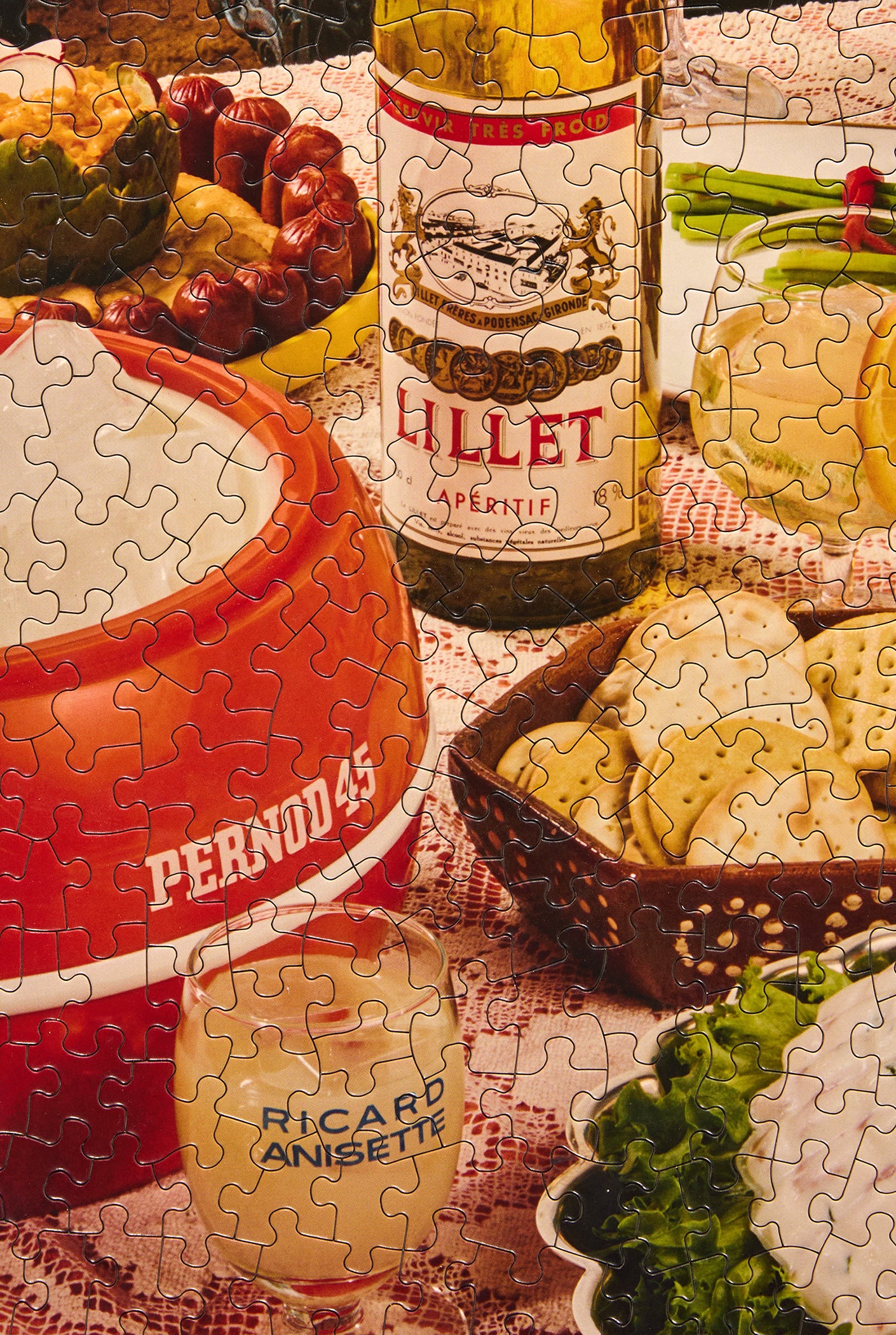 A jigsaw puzzle showing a cozy tabletop spread with a bottle of Lillet Caprirette liqueur, a red ceramic container labeled Perno 43, a small glass with 'Ricard Anisette' text, crackers, greens, and other food items.
