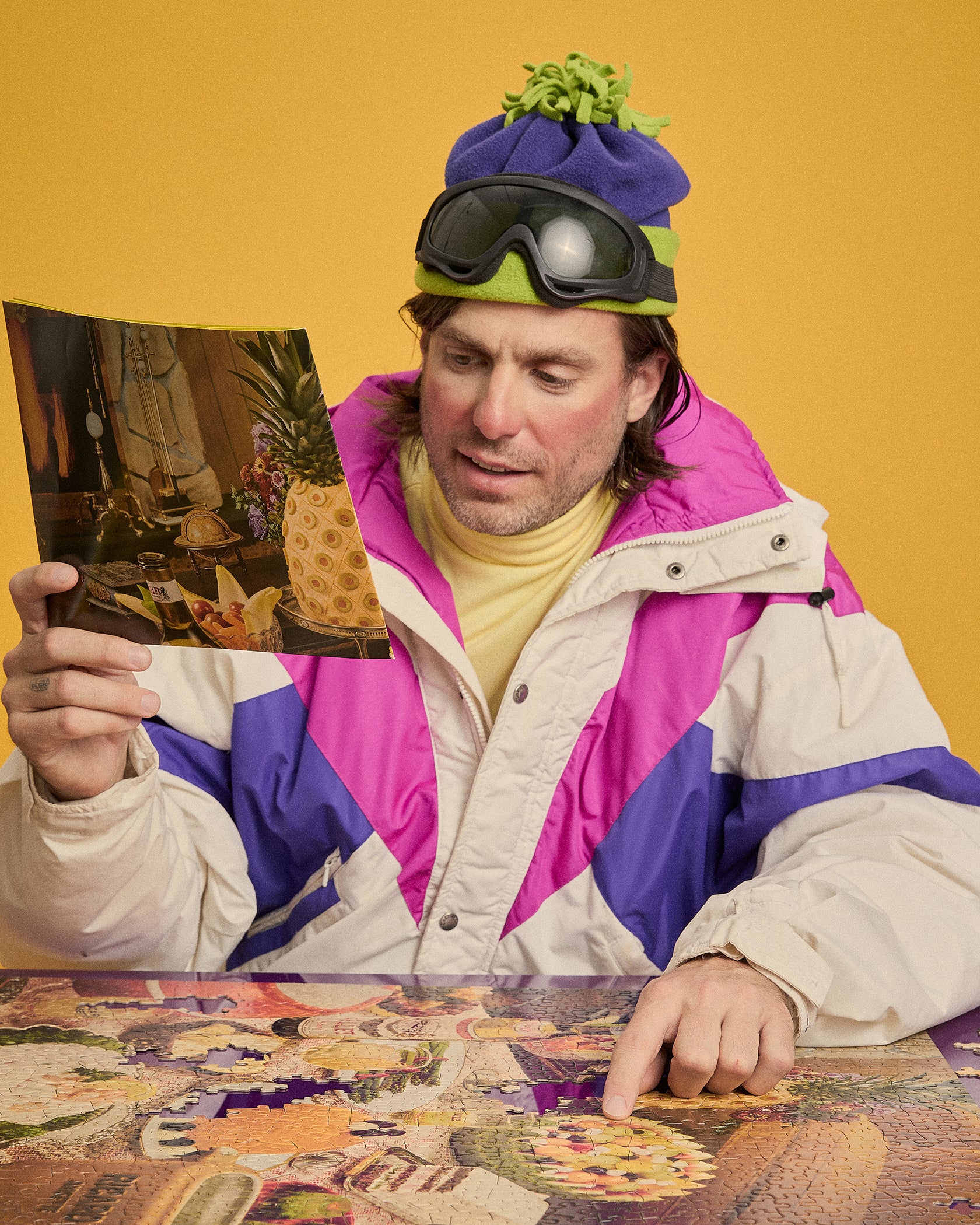 A man wearing a colorful vintage-style puffer jacket, a purple knit beanie with a green pom-pom, and ski goggles sits at a table against a mustard yellow background. He holds a picture in one hand and points to a detailed collage on the table with the other.