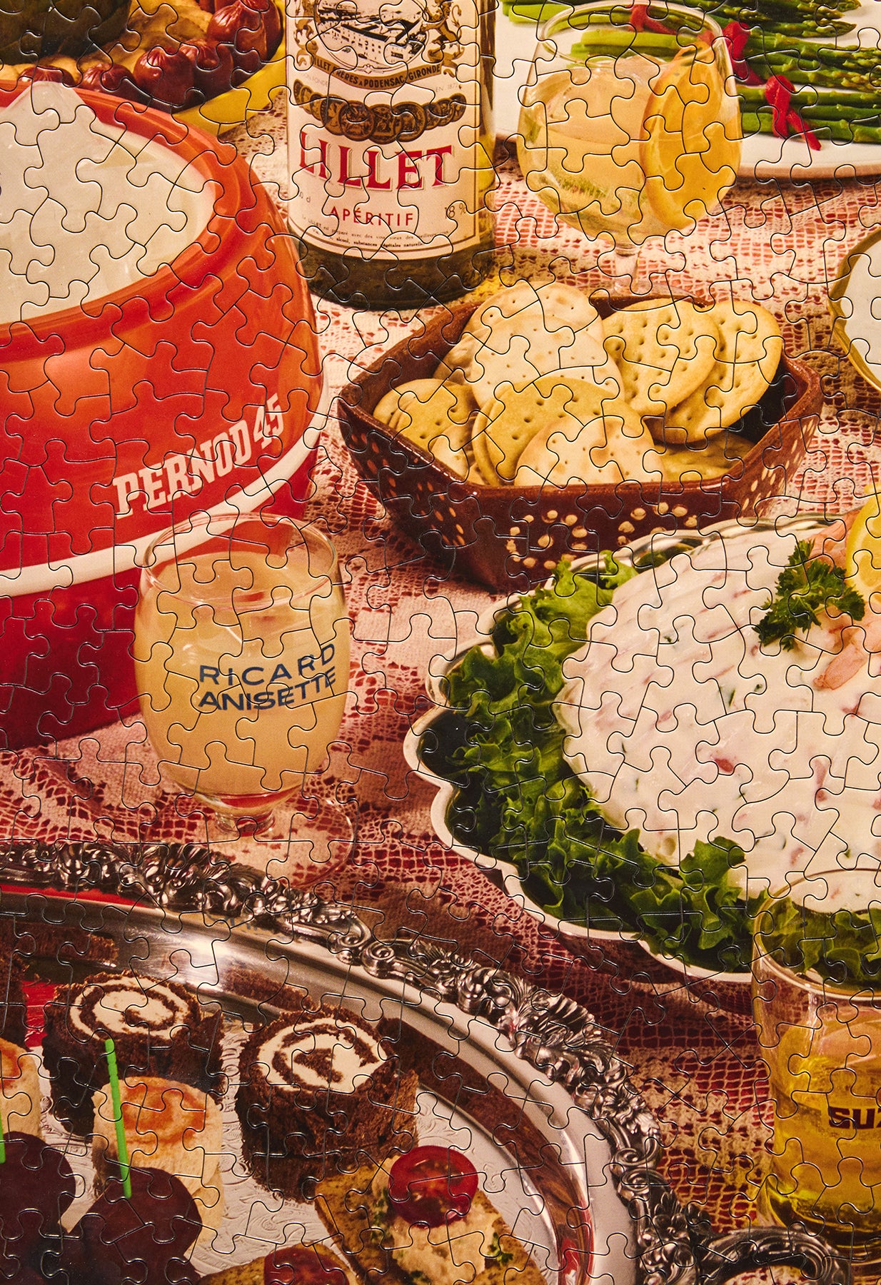 A jigsaw puzzle showing a cozy, rustic dinner table scene with food and drinks, including a red ceramic pot labeled 'PERNODAS', a bottle of Lillet, glasses, crackers, and plated dishes on a patterned tablecloth.