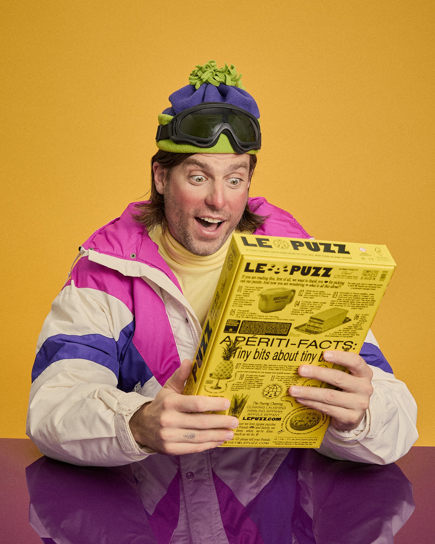 A man in a retro 1980s colorblock jacket, ski goggles, and beanie holds up a vintage-style yellow magazine or book titled 'LE PUZZ' against a solid yellow studio background.