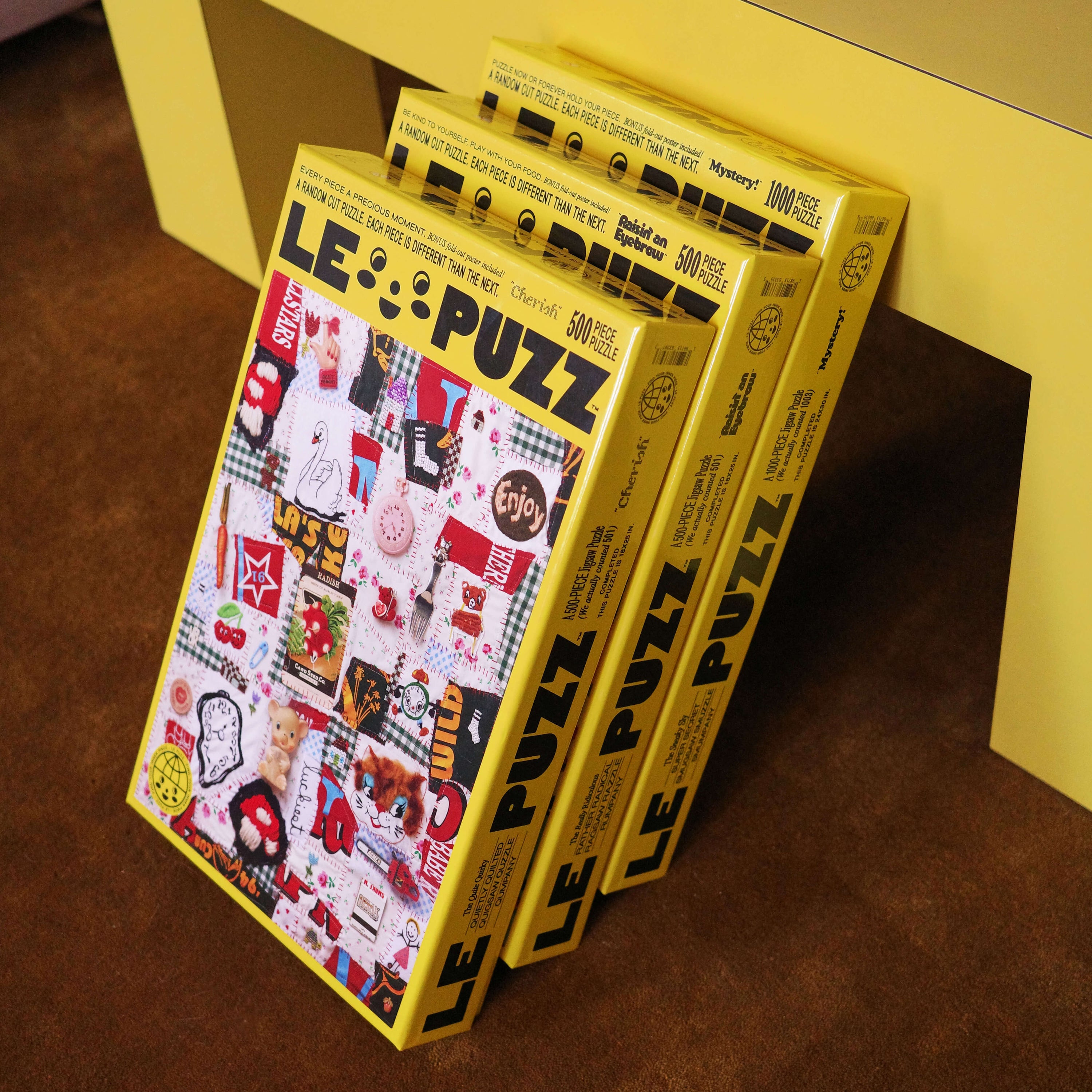 Three 'LE PUZZ' puzzle boxes stacked on a yellow surface.