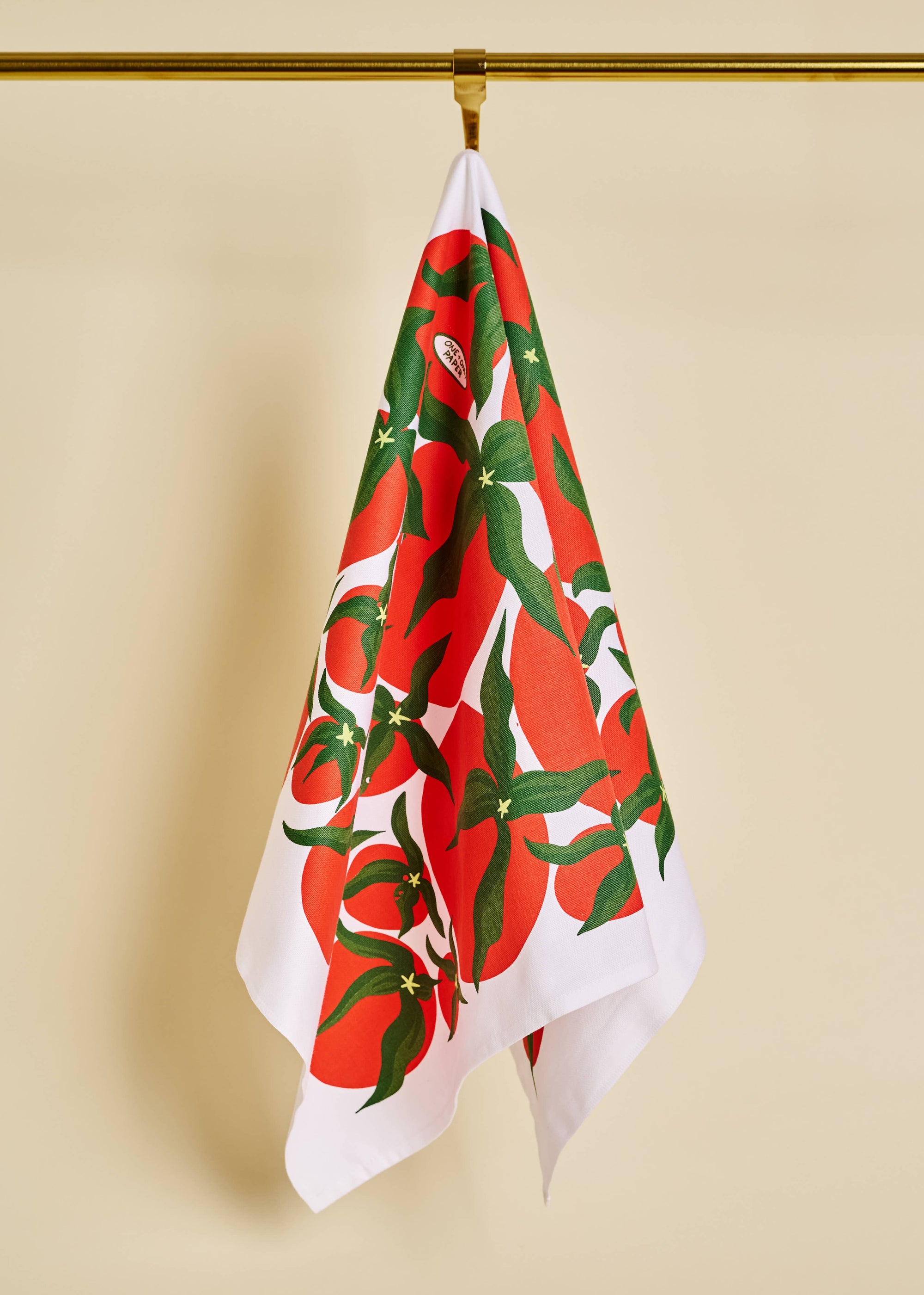 Tea towel with red and green tomato pattern hanging on a gold hook against a beige background