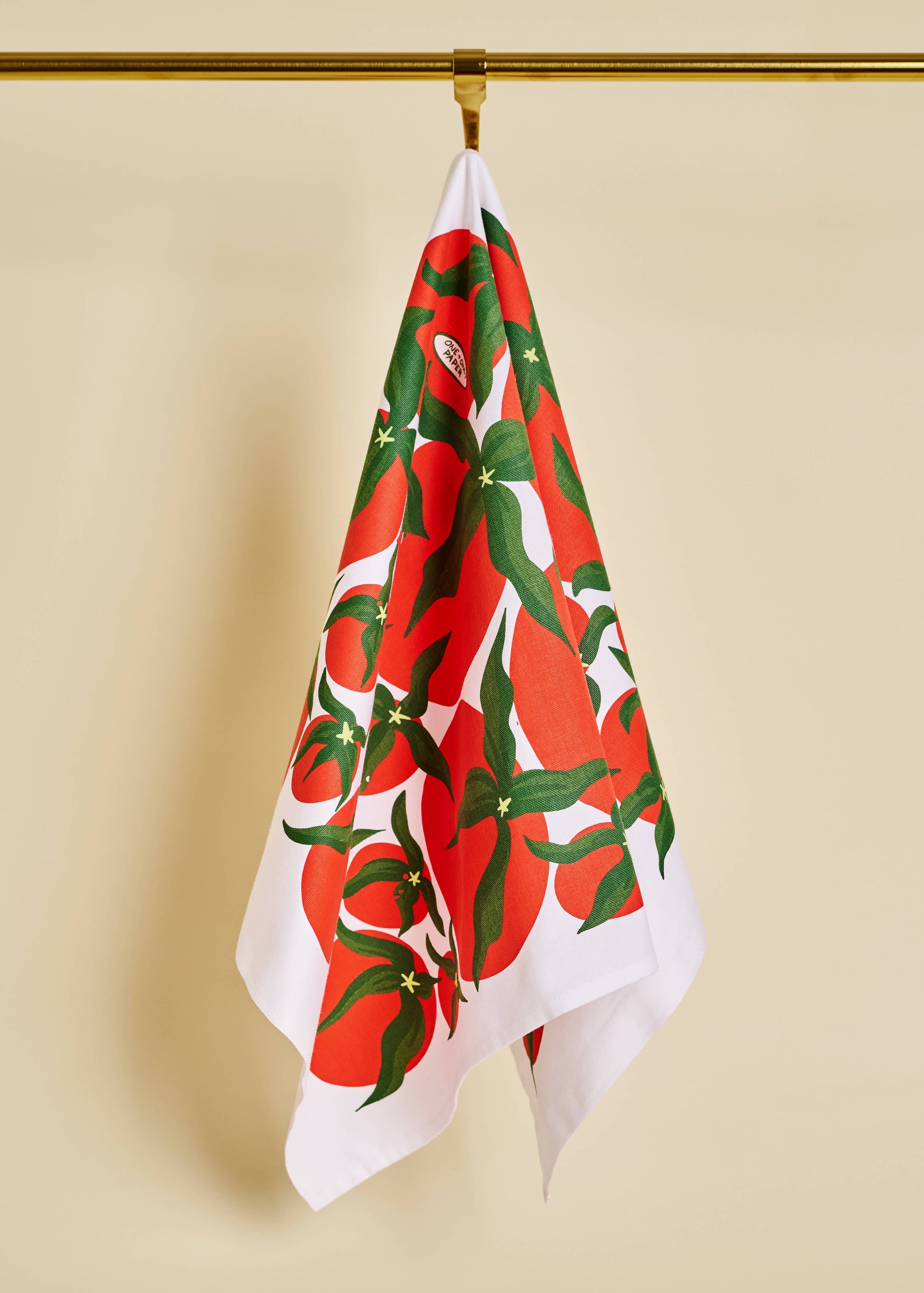Tea towel with red and green tomato pattern hanging on a gold hook against a beige background
