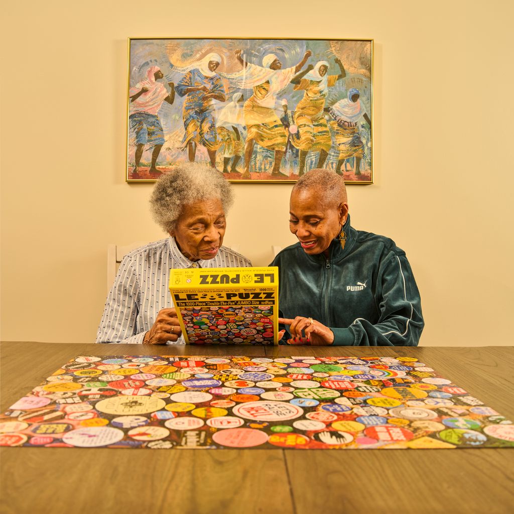 Two women sitting at a table with a colorful puzzle, a painting on the wall behind them.