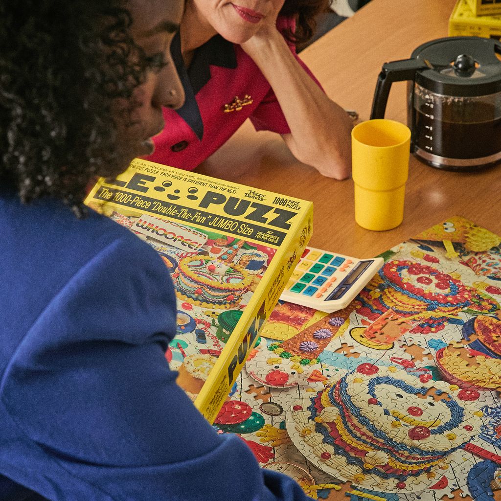 Two people working on a large puzzle with a colorful design, a yellow cup, and a coffee maker in the background.