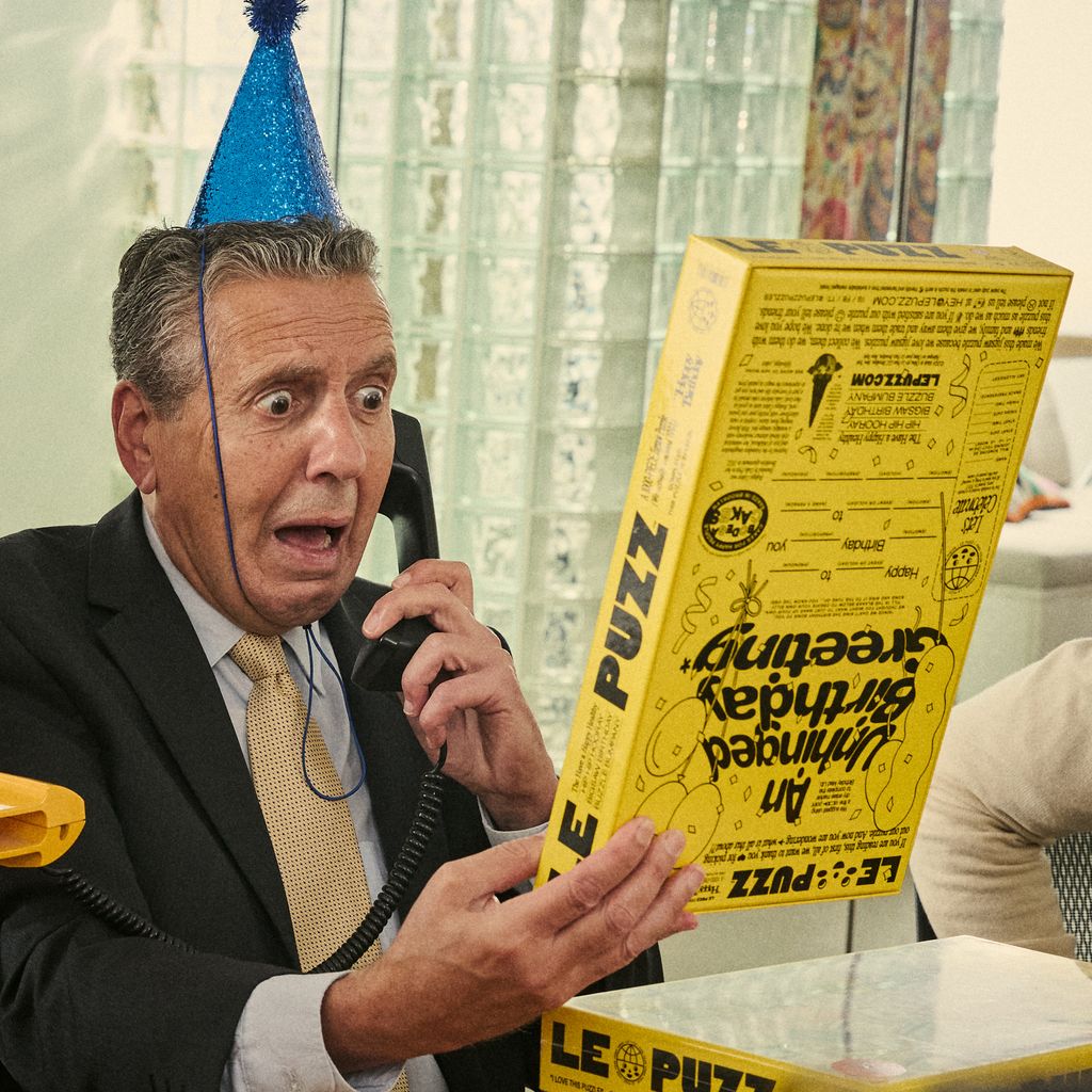 Man in a suit holding a yellow puzzle box and talking on the phone, wearing a blue party hat.