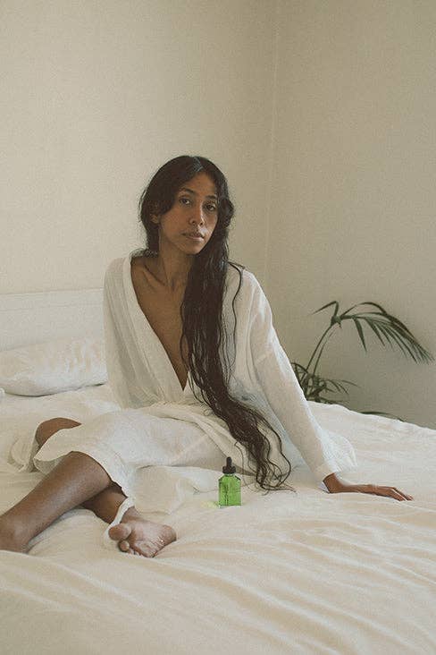 Woman in a white robe sitting on a bed with a bottle of green liquid.