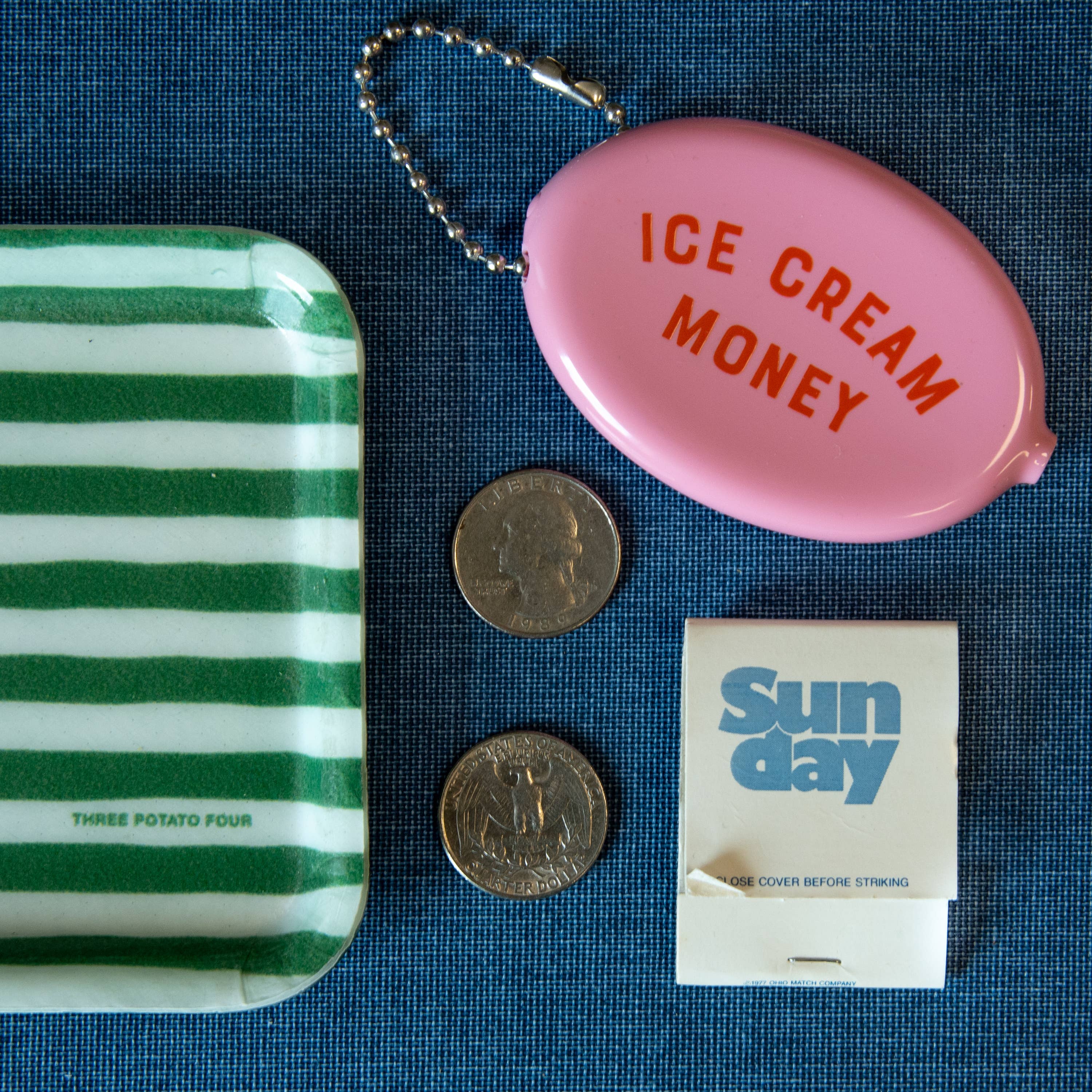 Pink coin purse with 'ICE CREAM MONEY' text, green striped tray, coins, and matchbook on a blue textured surface.