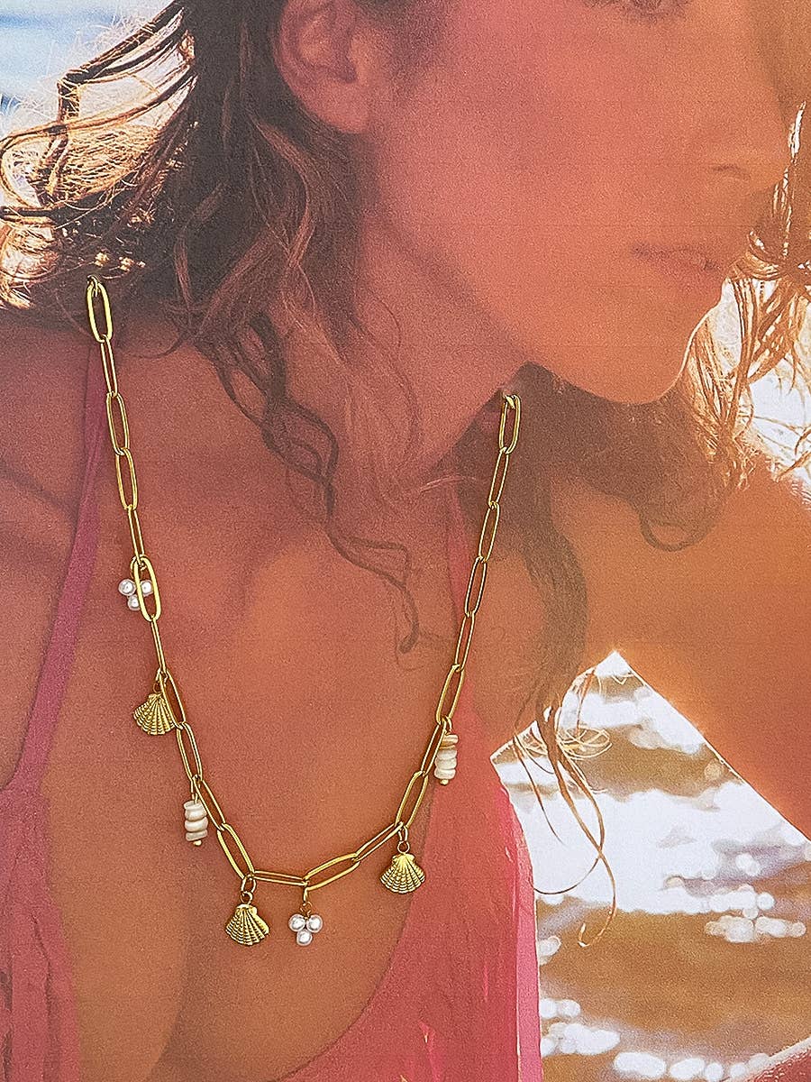 Woman wearing a gold chain necklace with shell and pearl charms on a beach.
