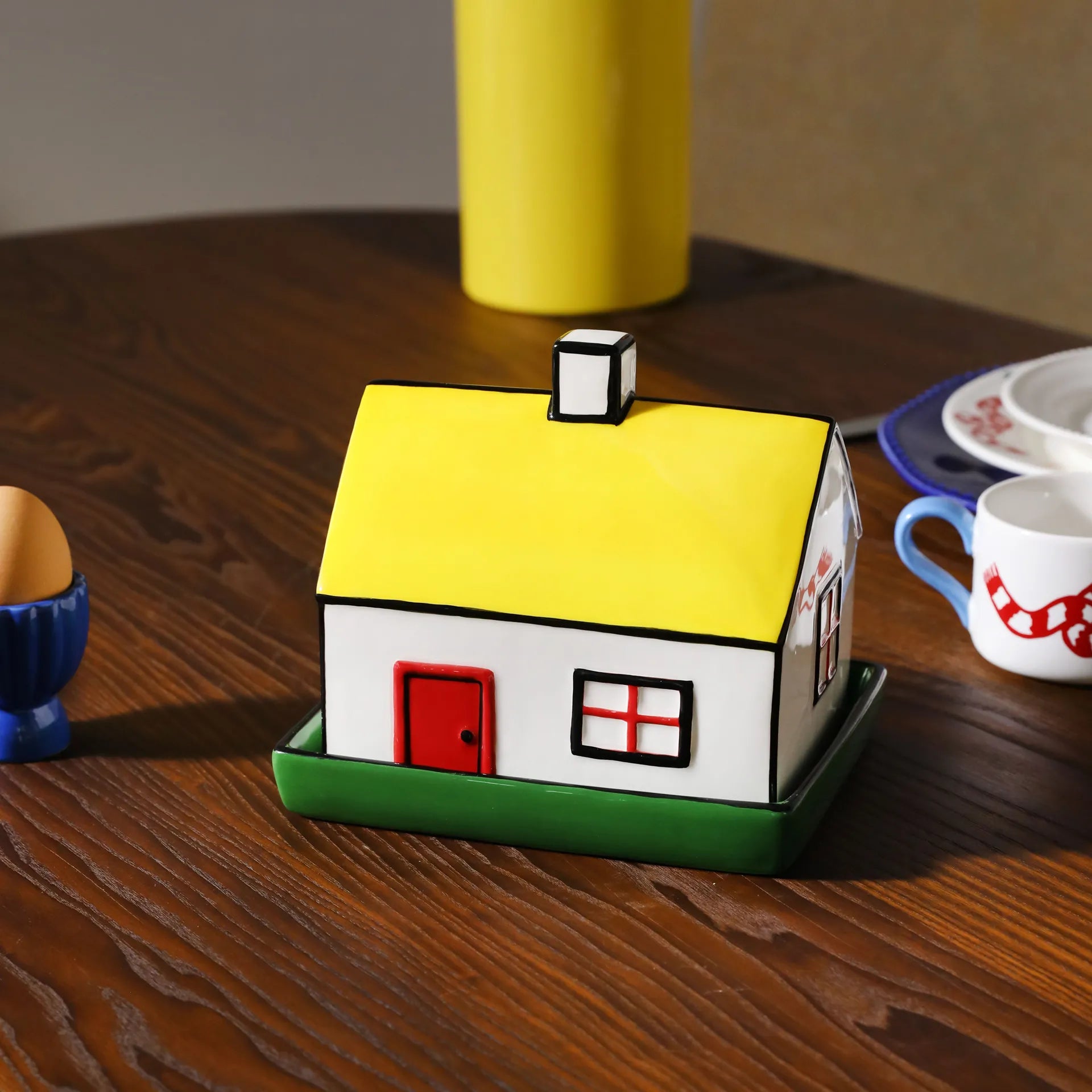 House-shaped butter dish on a wooden table with cups and a vase in the background
