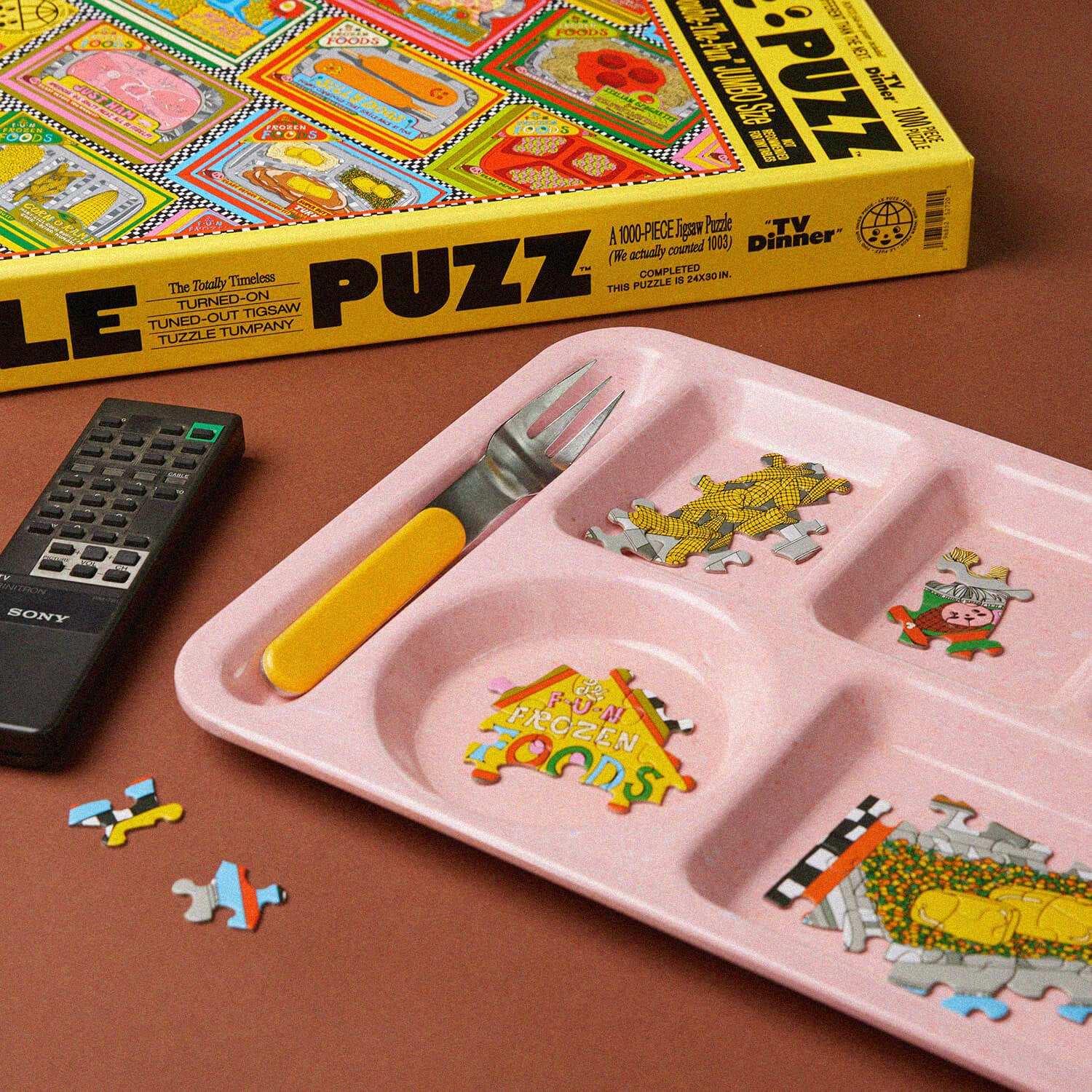 Pink children's tray with puzzle pieces, TV dinner box, and remote control on a brown surface