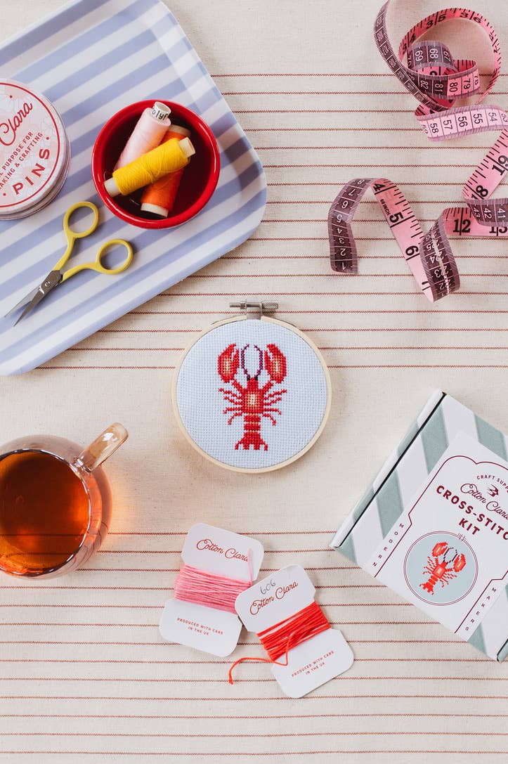 Cross-stitch kit with lobster design, thread, scissors, and tea on a wooden surface.