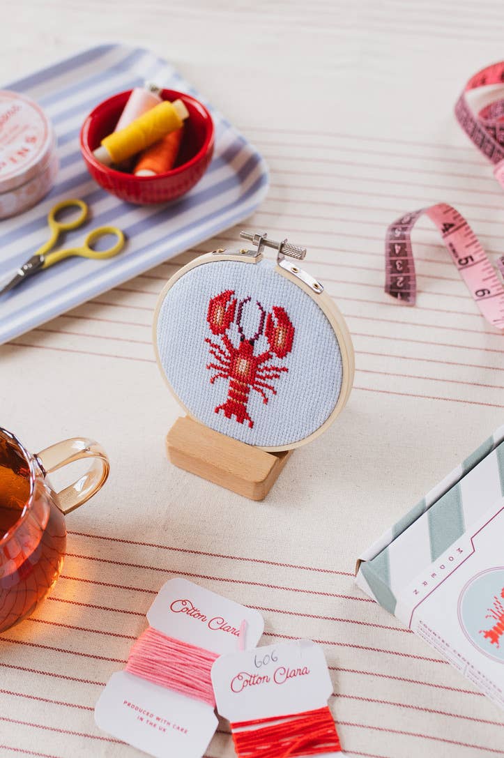 Cross-stitched lobster design in a hoop on a table with embroidery supplies.