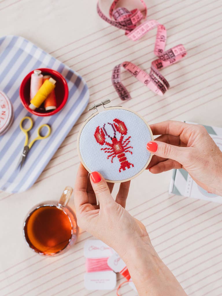 Person holding a cross-stitched lobster design on a small round fabric, with sewing supplies in the background.