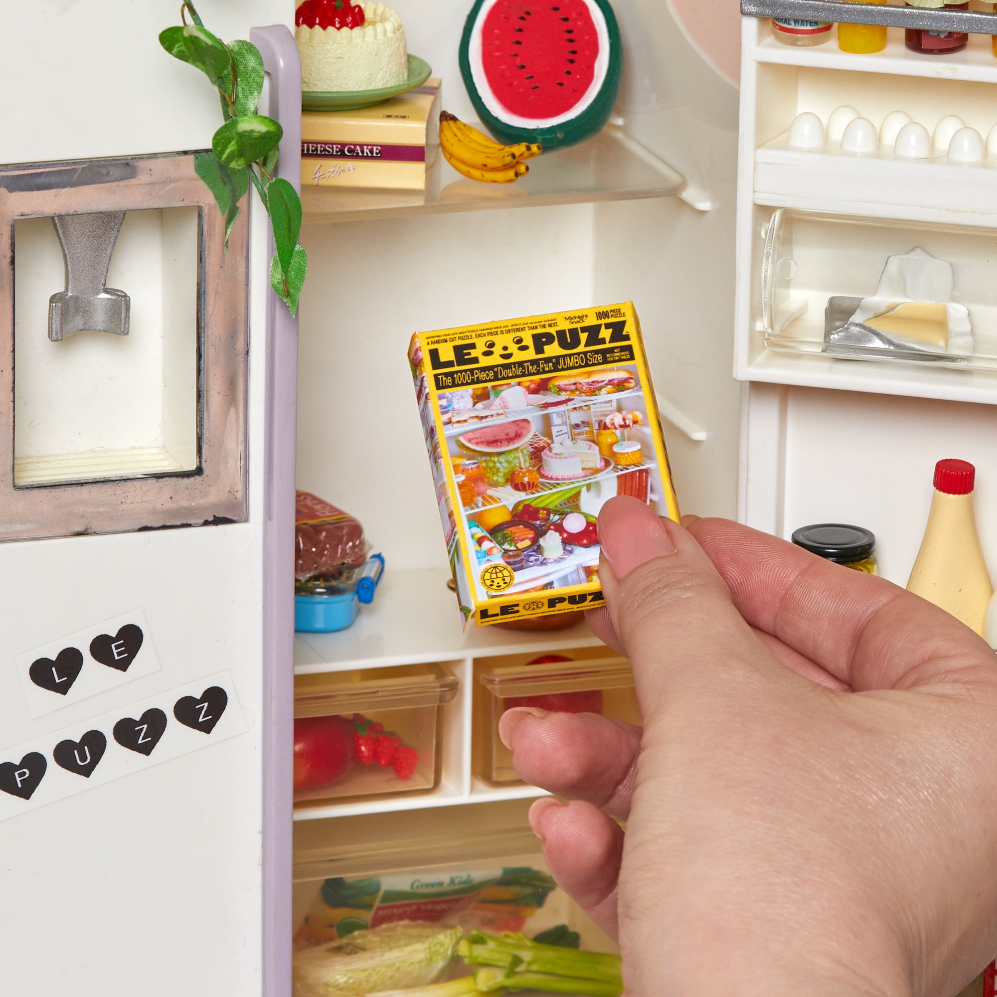 A hand holding a miniature toy box labeled 'LE PUZZ' inside a small doll's refrigerator with miniature food items and shelves.